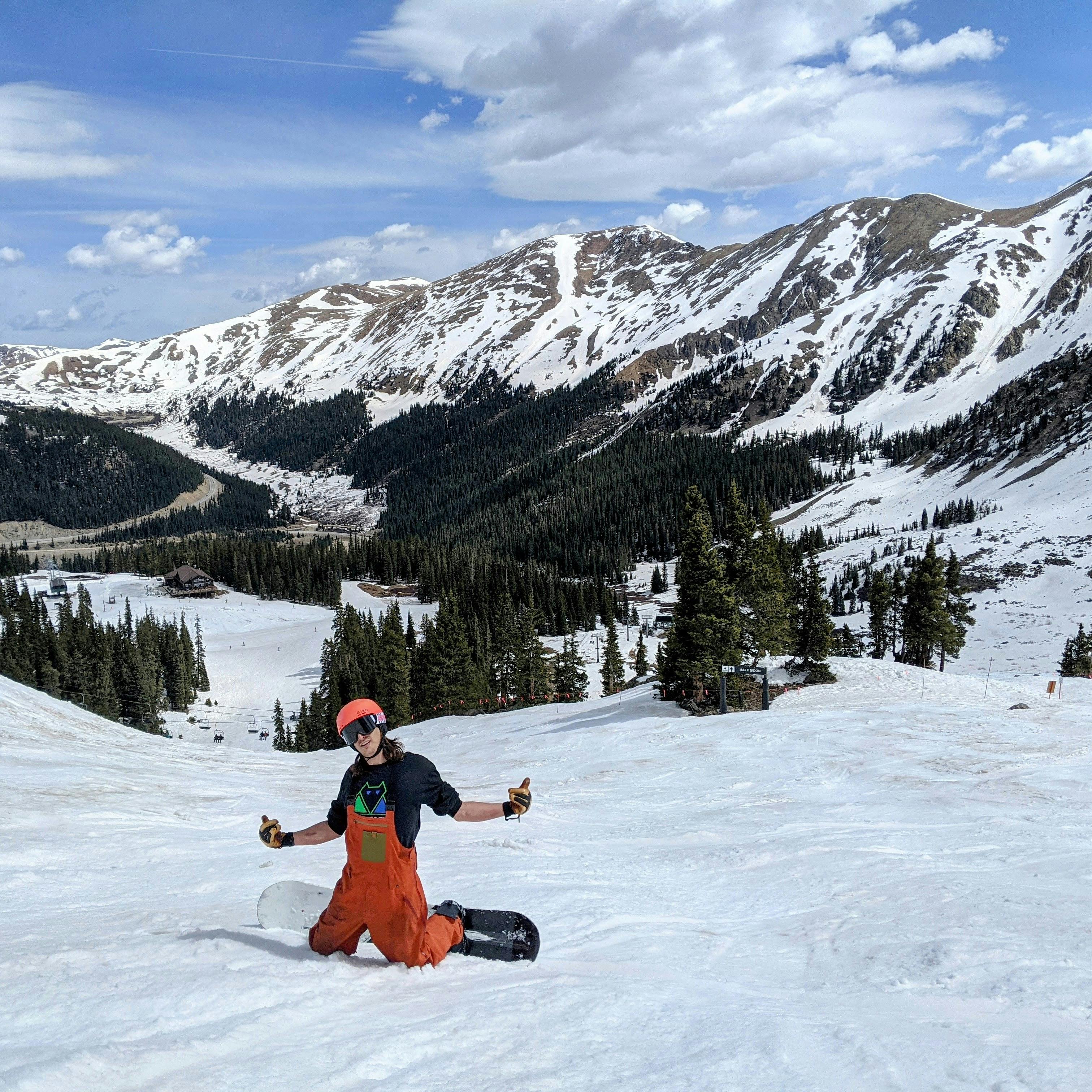 A snowboarder kneeling on a ski run with his arms up. There is a snowy mountain in the background. 