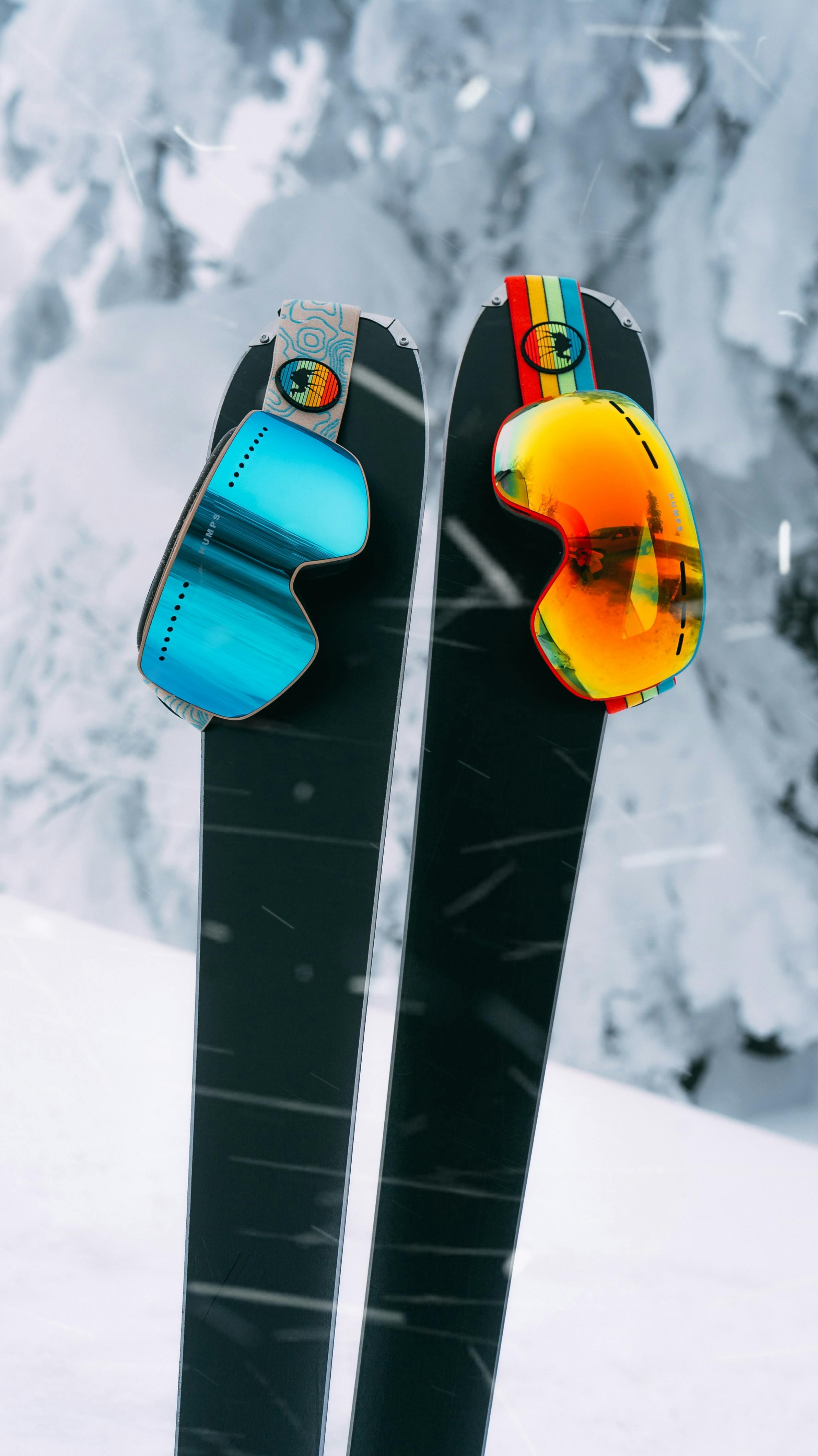 A pair of skis have ski googles balanced on them, one blue and one yellow tinted. 