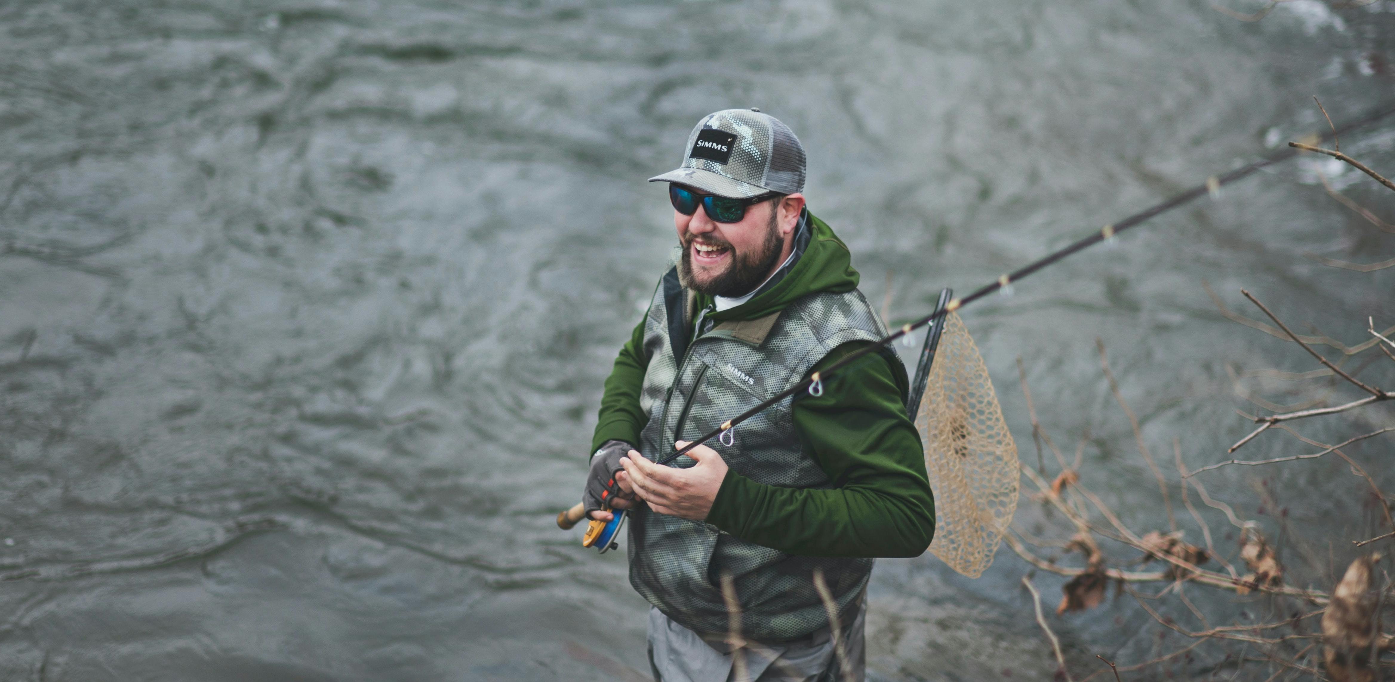 A man with fishing gear and wearing sunglasses stands in a river holding his fly rod.