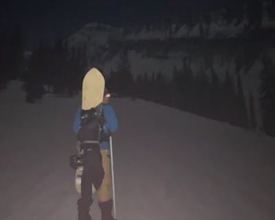 A snowboarder walking in the dark with the Nidecker Mellow Snowboard · 2022 on his back. 