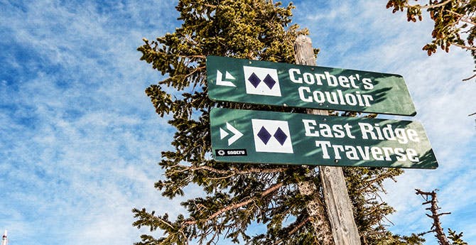 Two signs on a post read "Corbet's Couloir" with a double black diamond and "East Ridge Traverse" with a double black diamond.