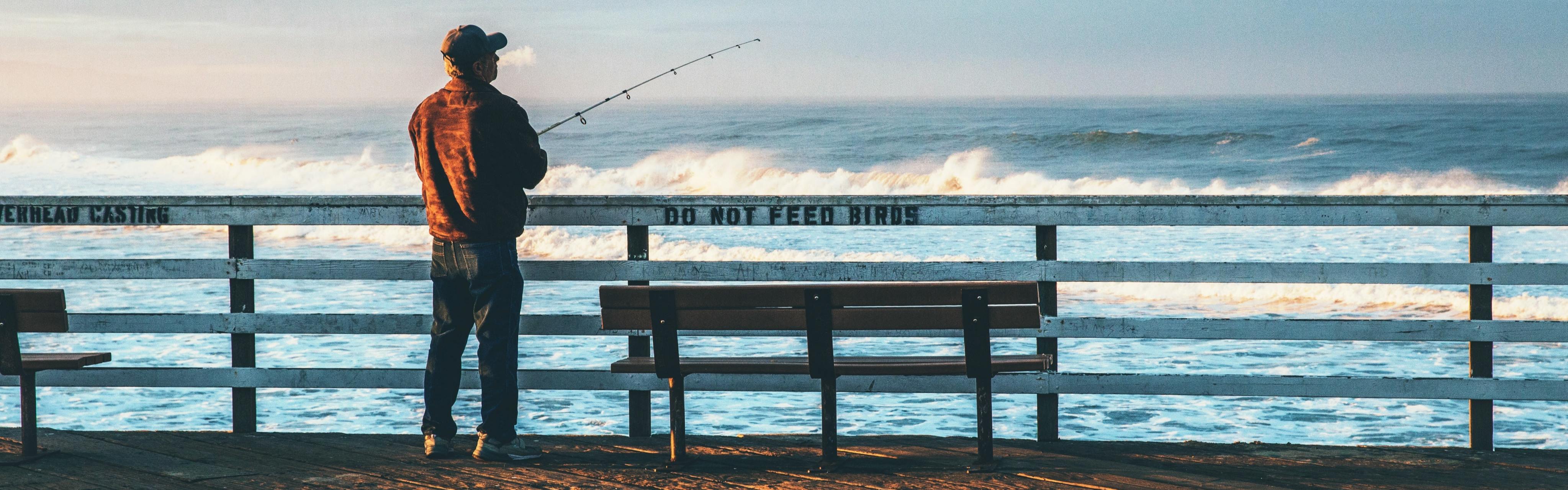 A man fishes off of a pier with waves crashing in the background. Painted in black on the white railing of the pier is "DO NOT FEED BIRDS."
