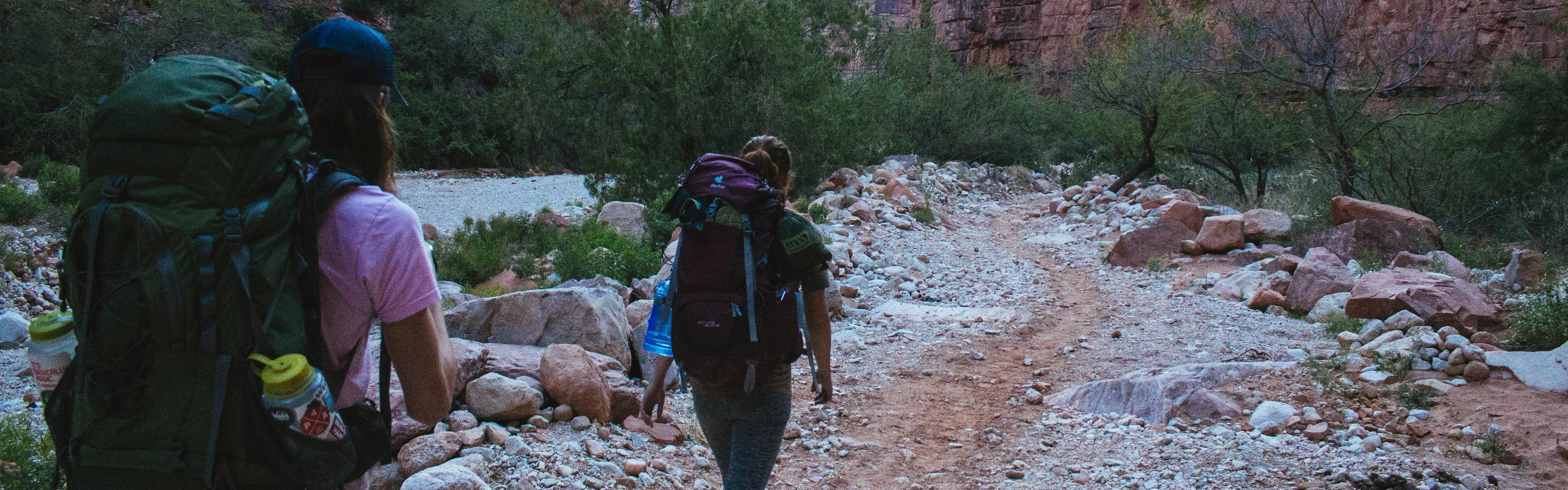 Two women walk down a rocky trail with large packs on their backs