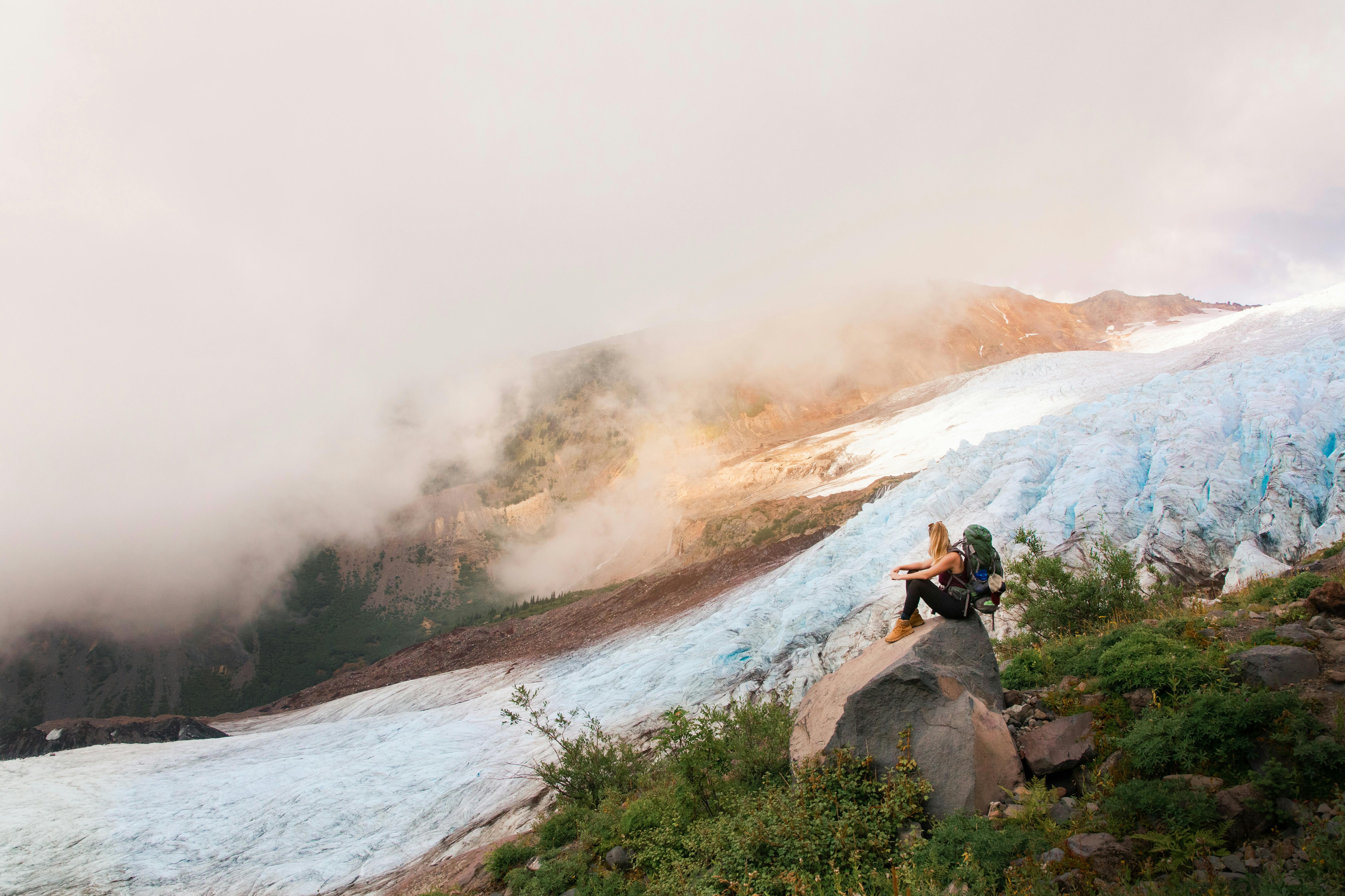 A woman sits on a boulder with her backpack on and looks out over a large snowfield. In the background, fog rolls down the hill. 