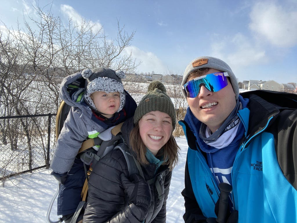 A family on a ski trail. There is a baby in the mothers backpack and the father is wearing sunglasses.