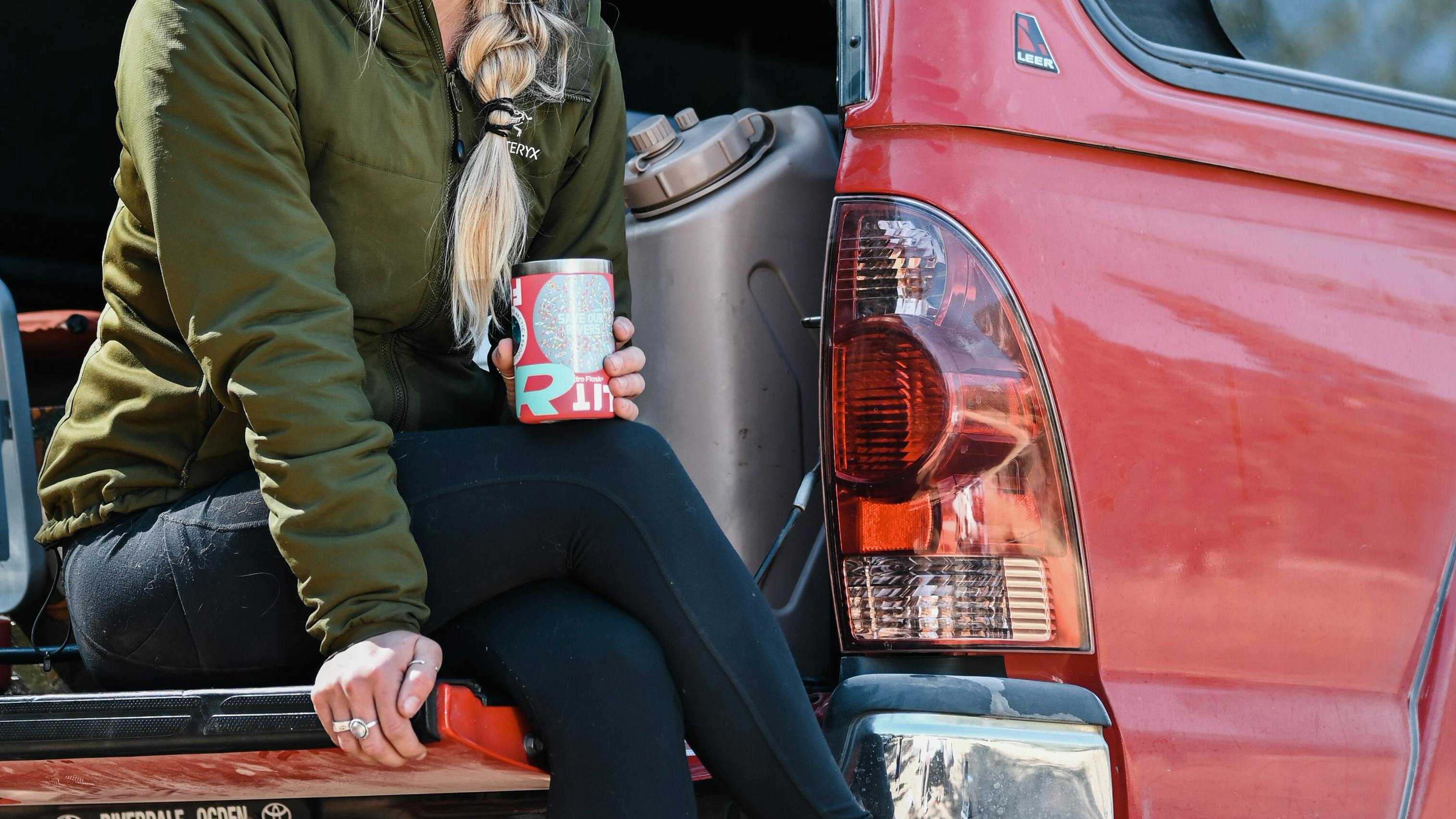 A woman sitting on the back of a truck with a stove out, a water jug in the back, and holding a HydroFlask 12 oz Cooler Cup.