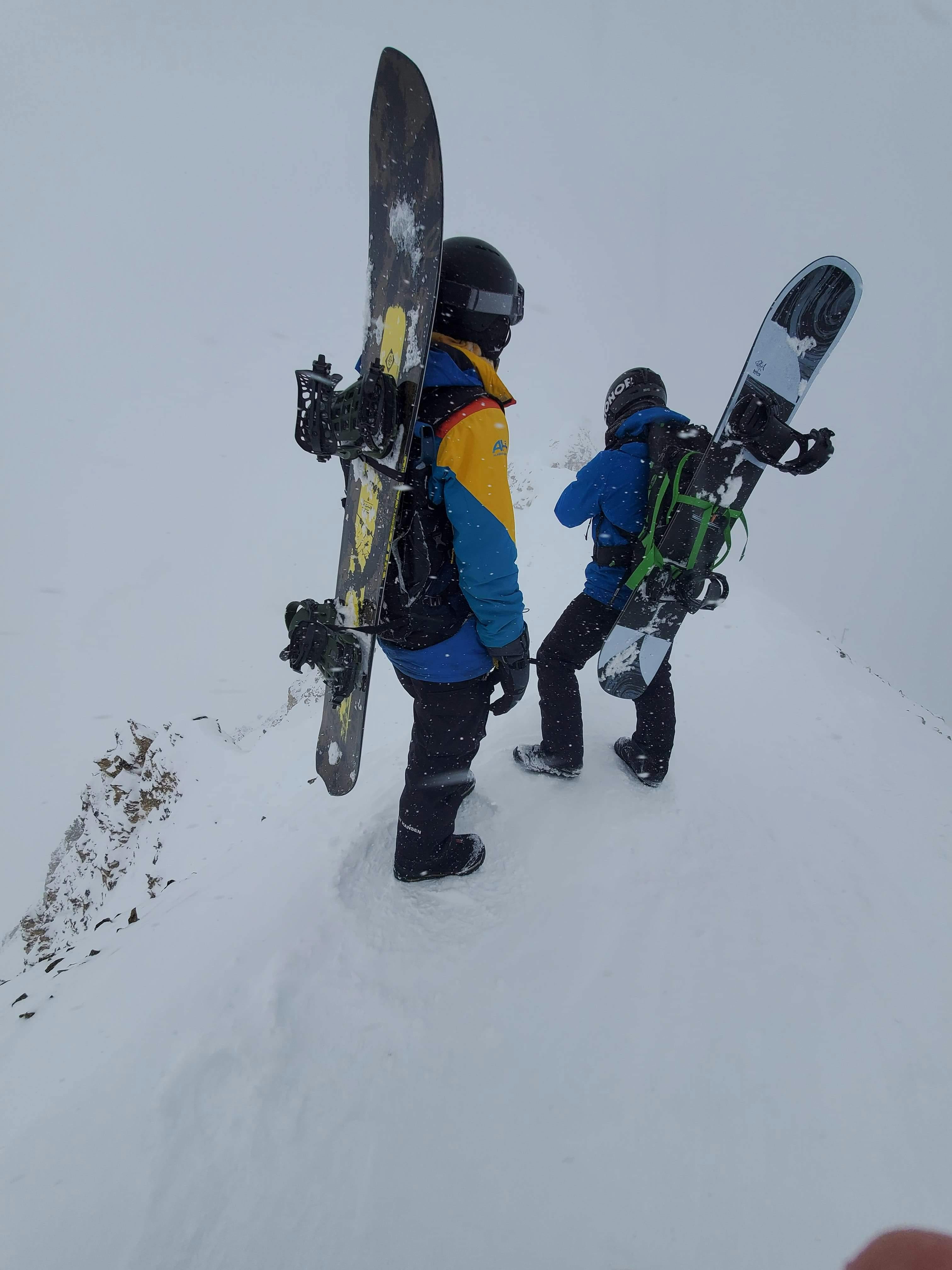 Two snowboarders wearing their snowboards on their back and standing at the top of a peak. 
