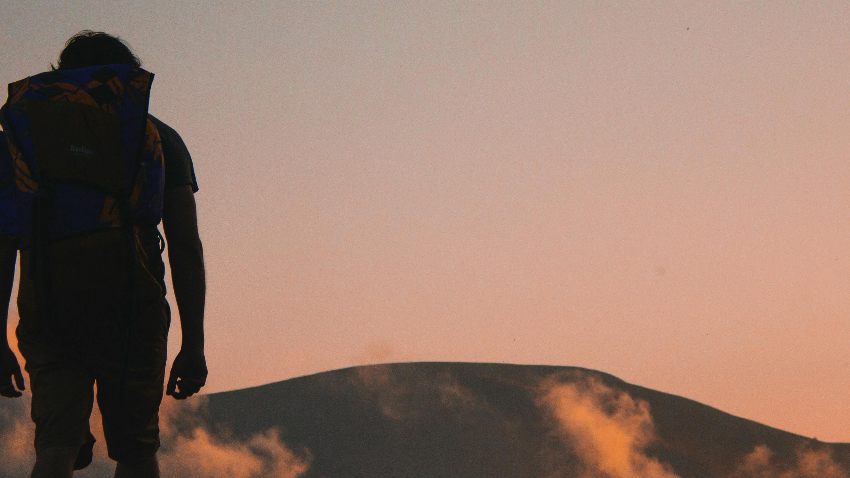 A person with a backpack is silhouetted from behind with sunset hills in front of him.