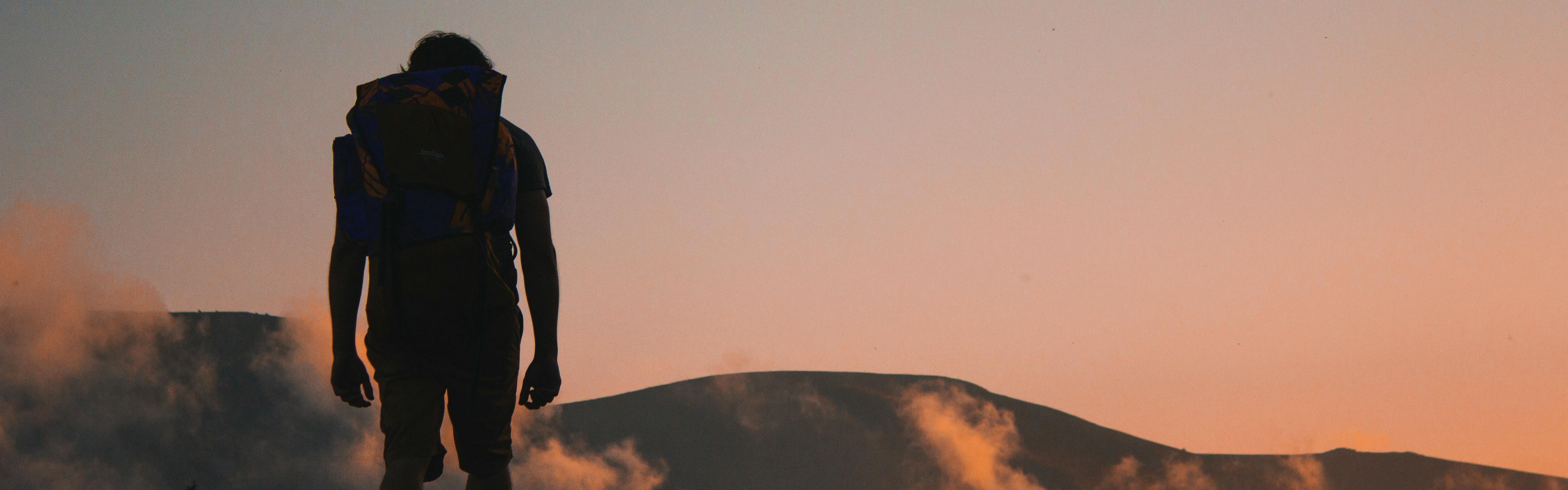 A person with a backpack is silhouetted from behind with sunset hills in front of him.