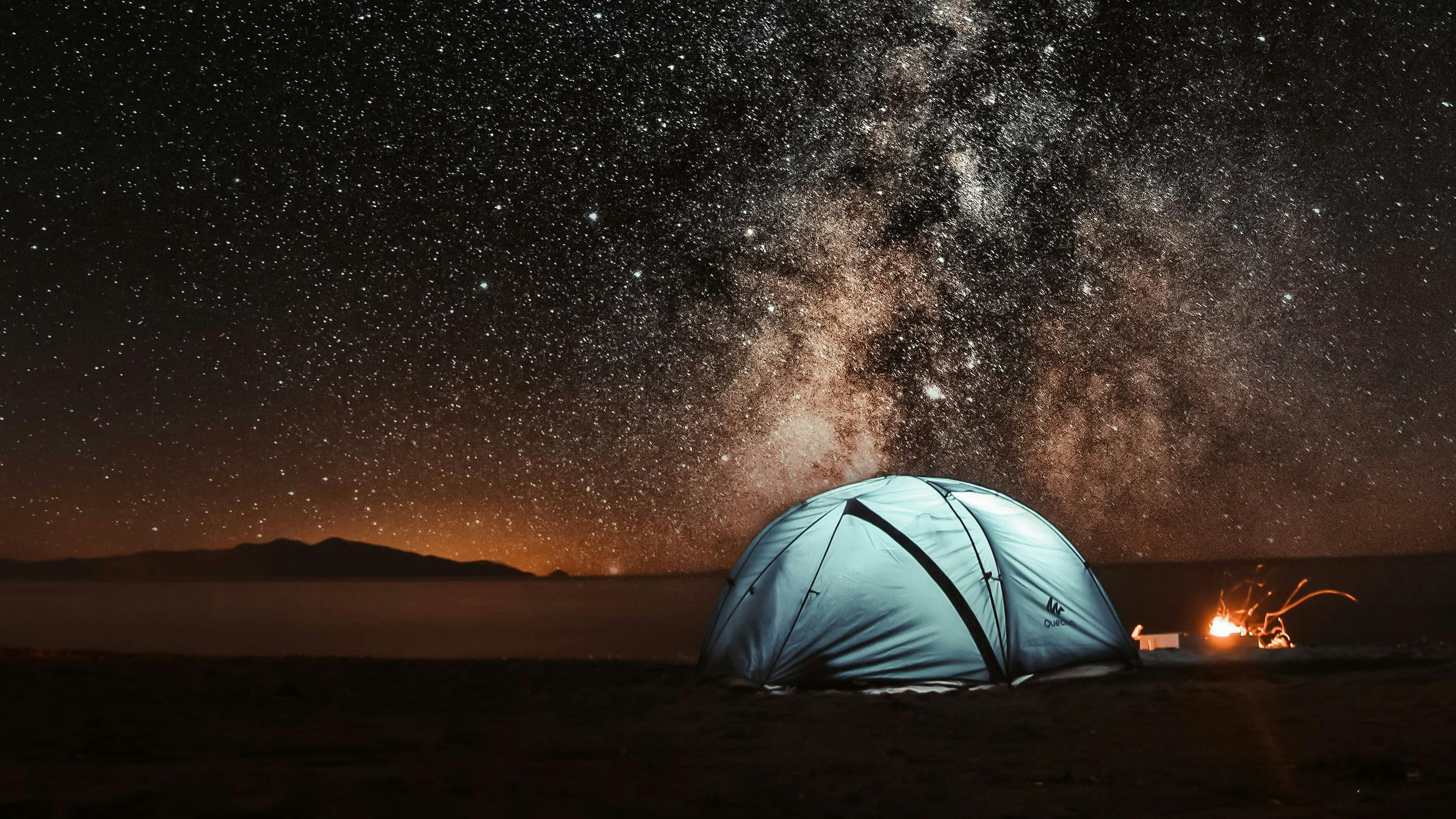 A brightly lit up tent sits in the wilderness against a starry sky