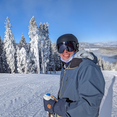 A skier standing on a ski hill. He is smiling at the camera and wearing the Flylow Men's Quantum Pro Shell Jacket. There are snowy trees in the background. 