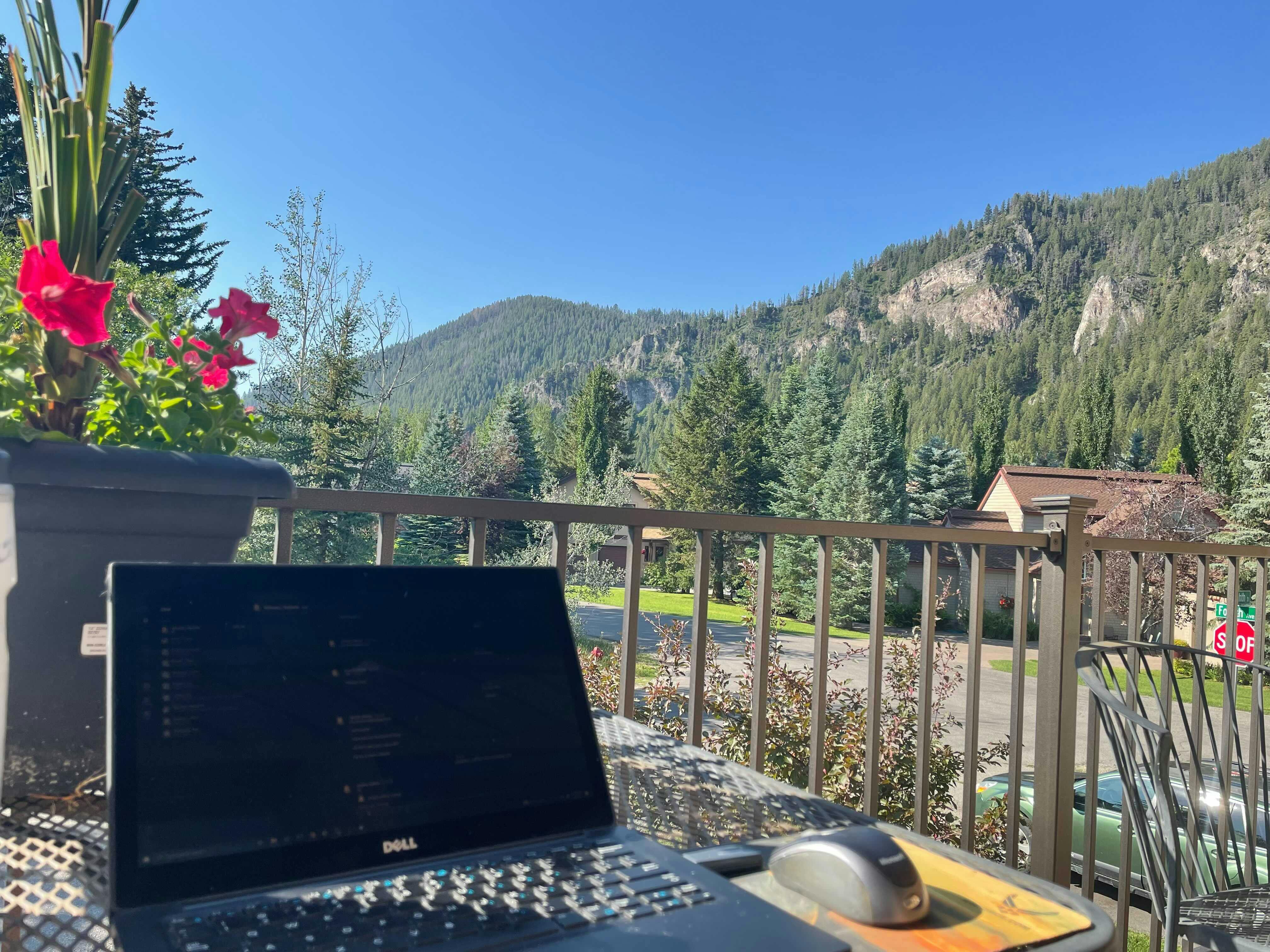 A laptop sits open on an outdoor table on a balcony in front of a mountain view. The sky is blue and the scene is sunny.