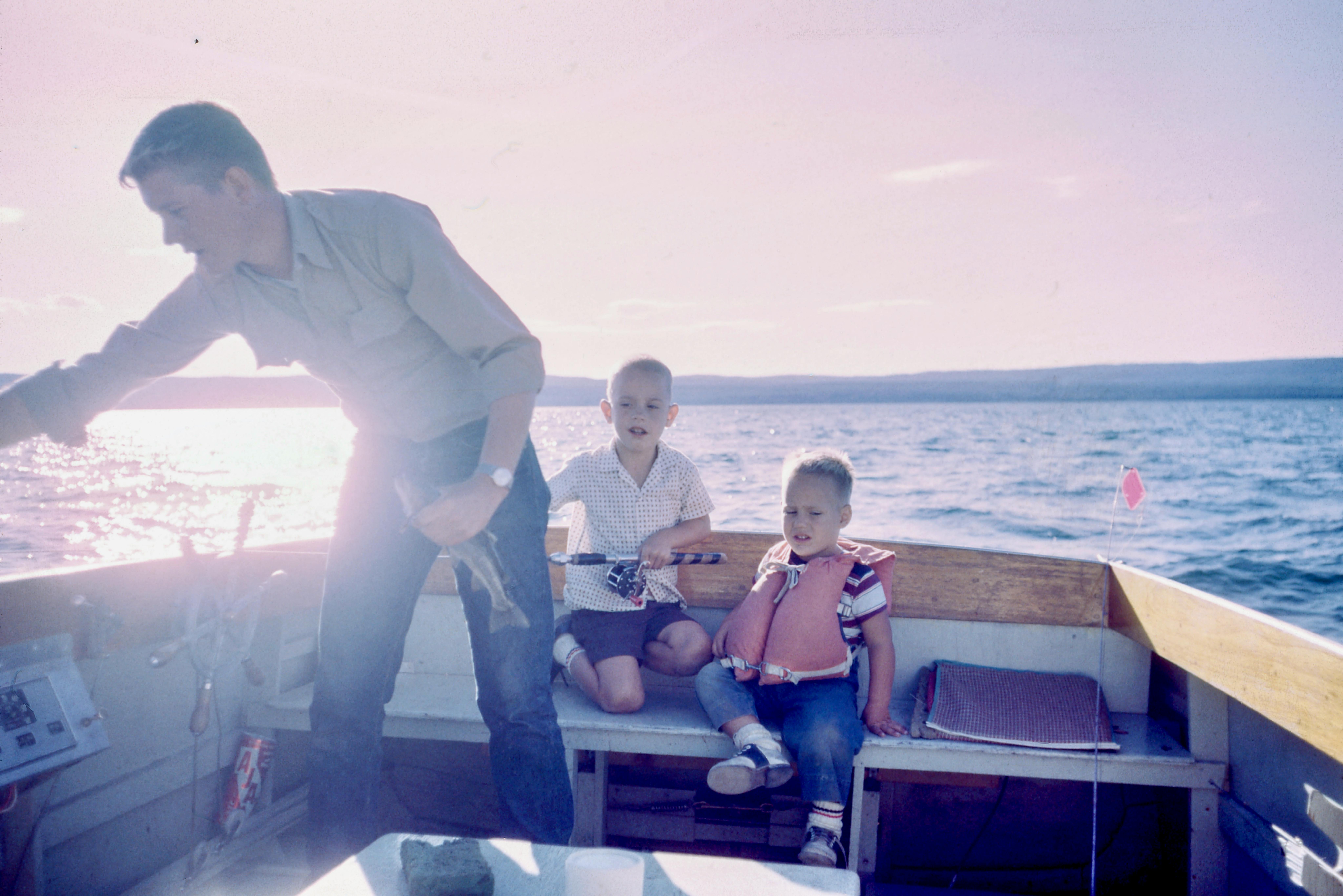 A man stands on a boat, holding a fish in one hand and leaning over to grab something with the other. Two young children, one in a life jacket, sit on a bench at the back of the boat and watch. 
