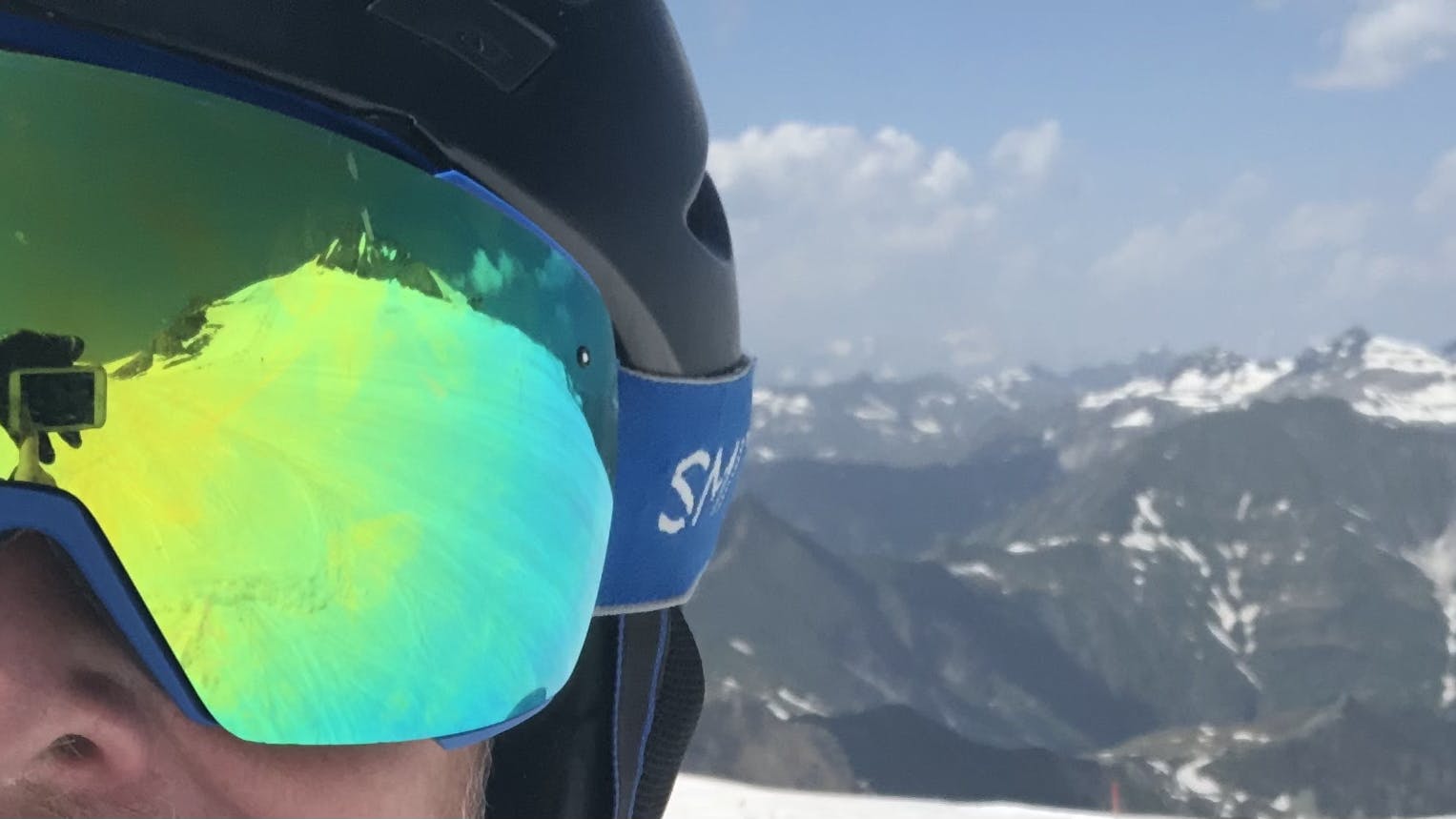 A man in a ski helmet and goggles takes a selfie on the ski slope. He is wearing the Smith I/O Mag goggles with the Sun Green Mirror Lens.