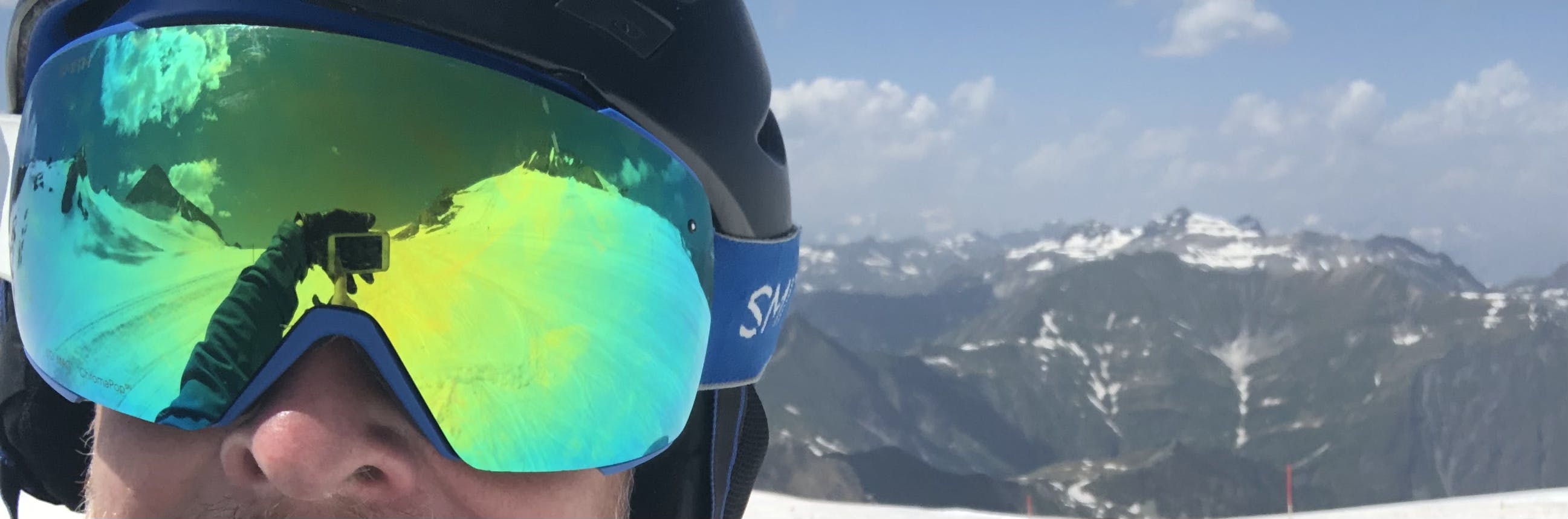 A man takes a selfie at a ski area while he is wearing goggles and a helmet. 