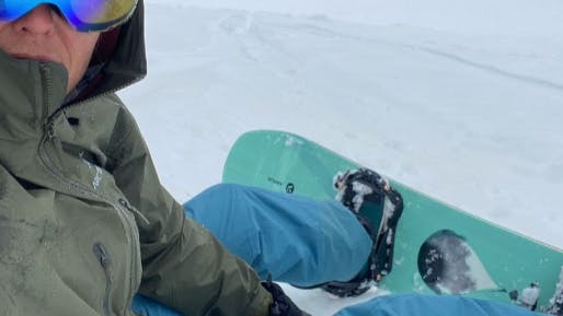A snowboarder taking a selfie of herself sitting on the snow with a snowboard on her feet.