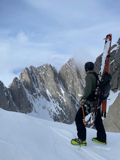 A man in ski gear stands at the top of a mountain pass looking out. He has skis strapped to his backpack and there are snowy mountains in the distance. 