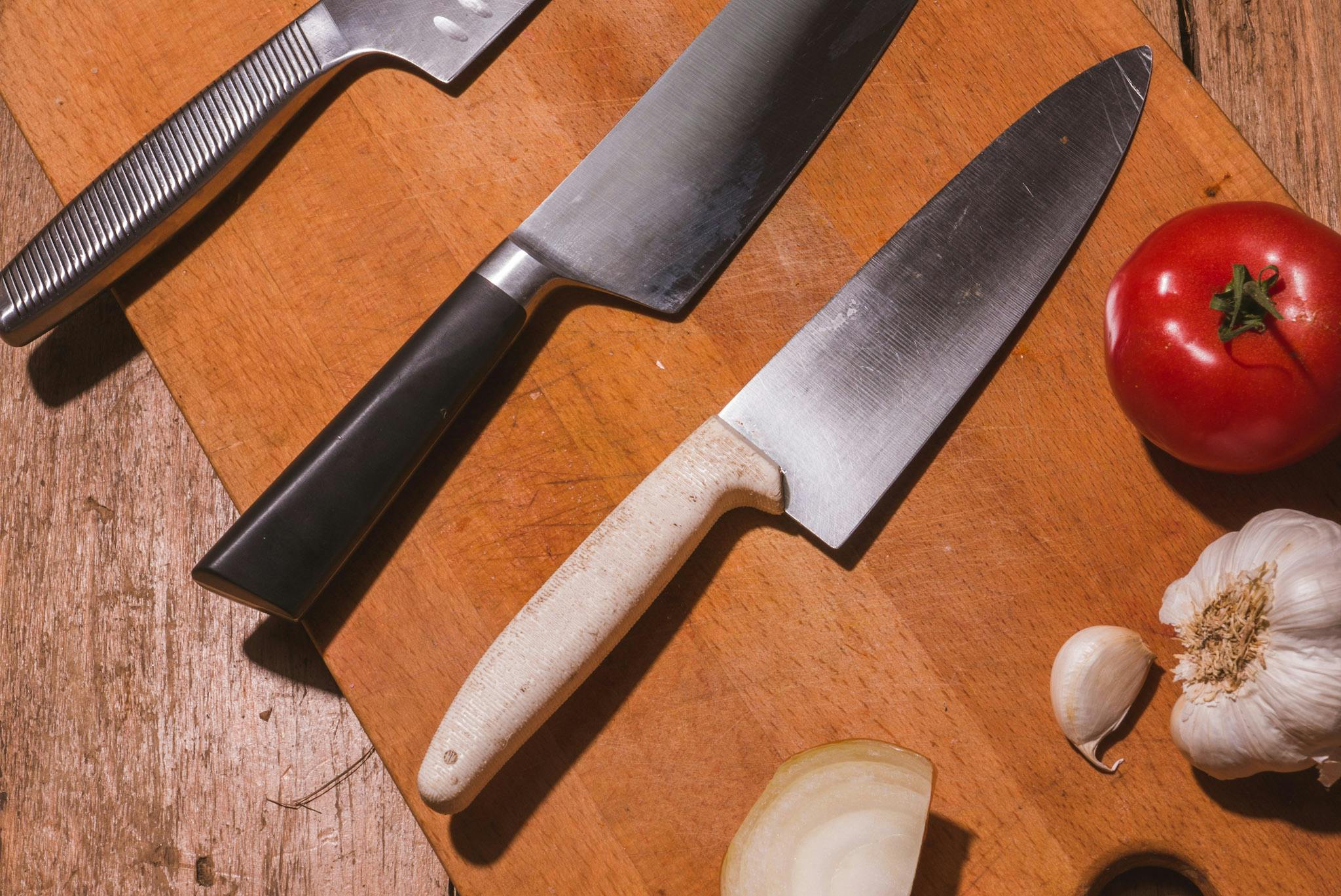 Three knives sitting on a cutting board with a tomato and garlic. 