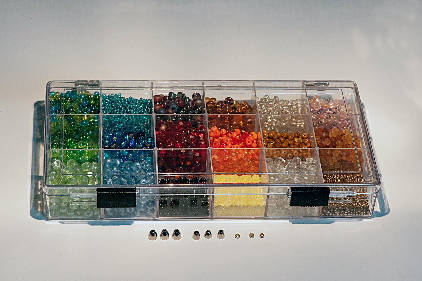 A container of beads for tying flies. There are several colored beads in blues, greens, oranges, and browns. There are a few silver ones sitting outside of the box in different sizes.
