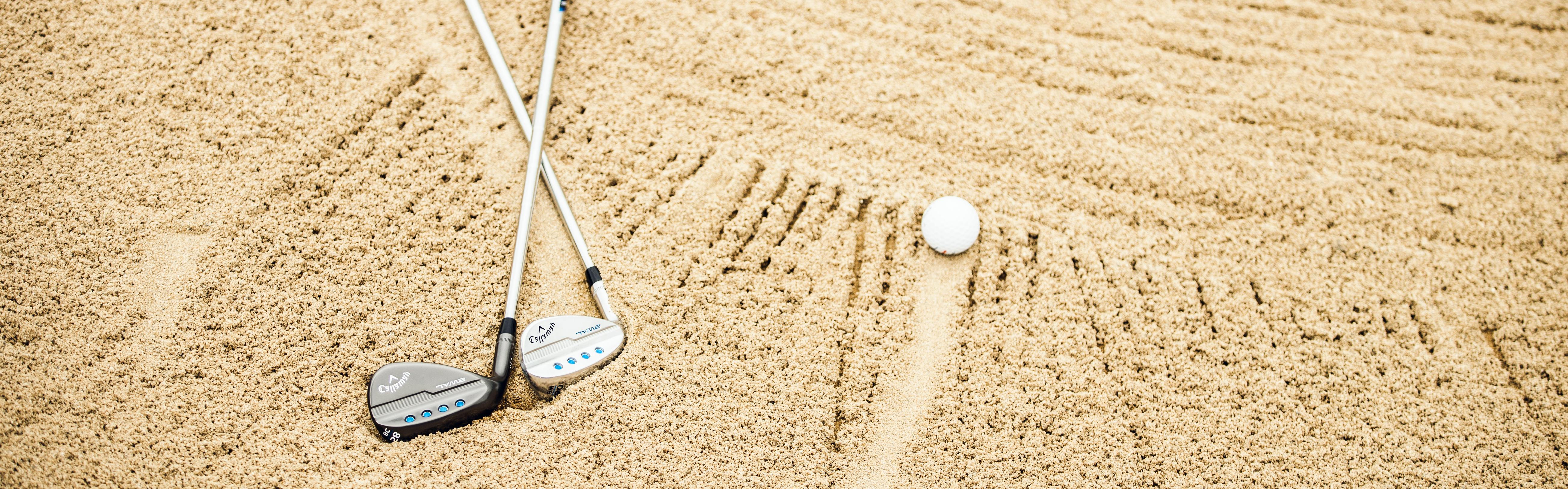 Two golf wedges in the sand next to a golf ball