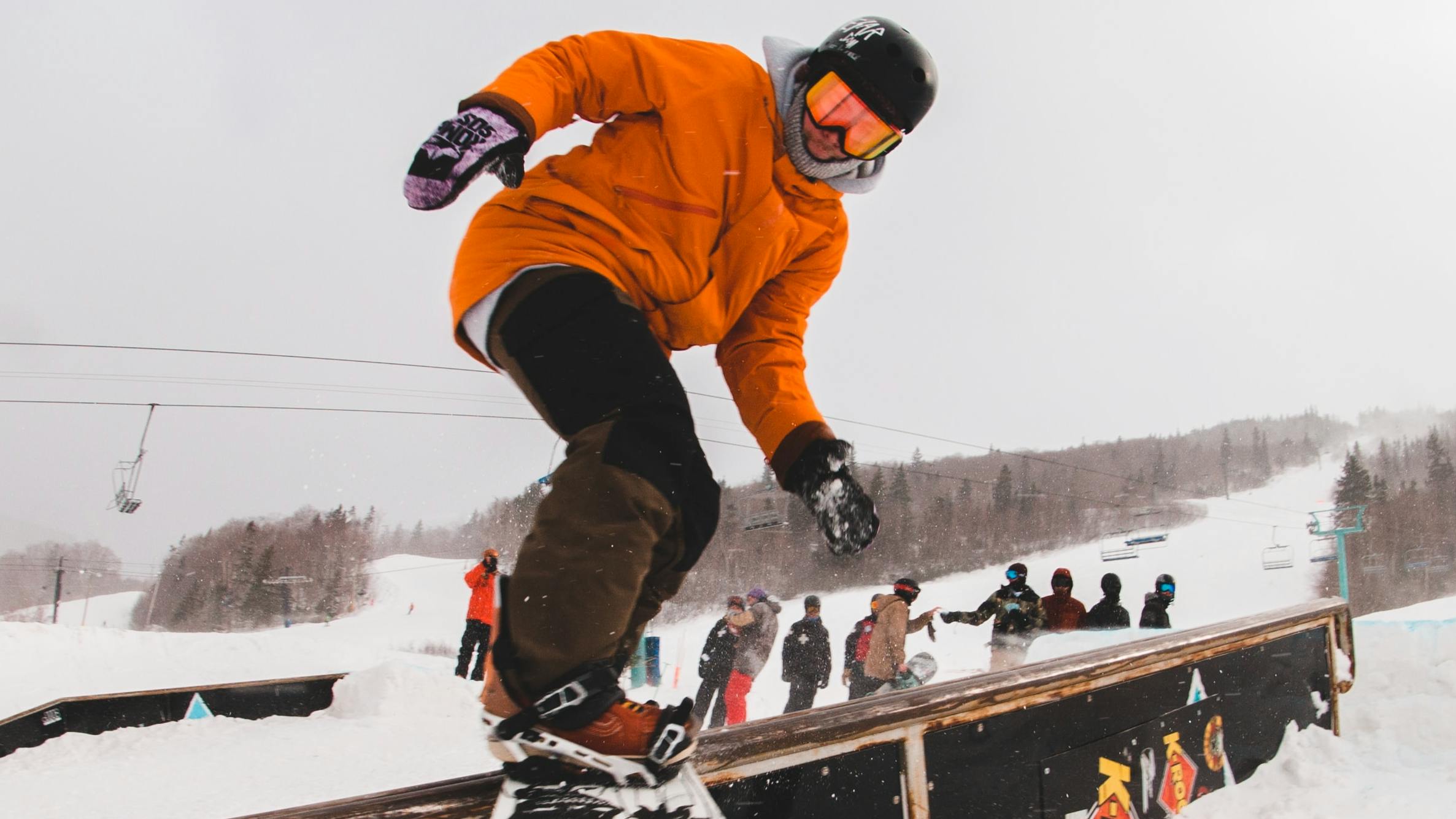 Snowboarder wearing orange jacket slides across a rail on his snowboard in the terrain park.
