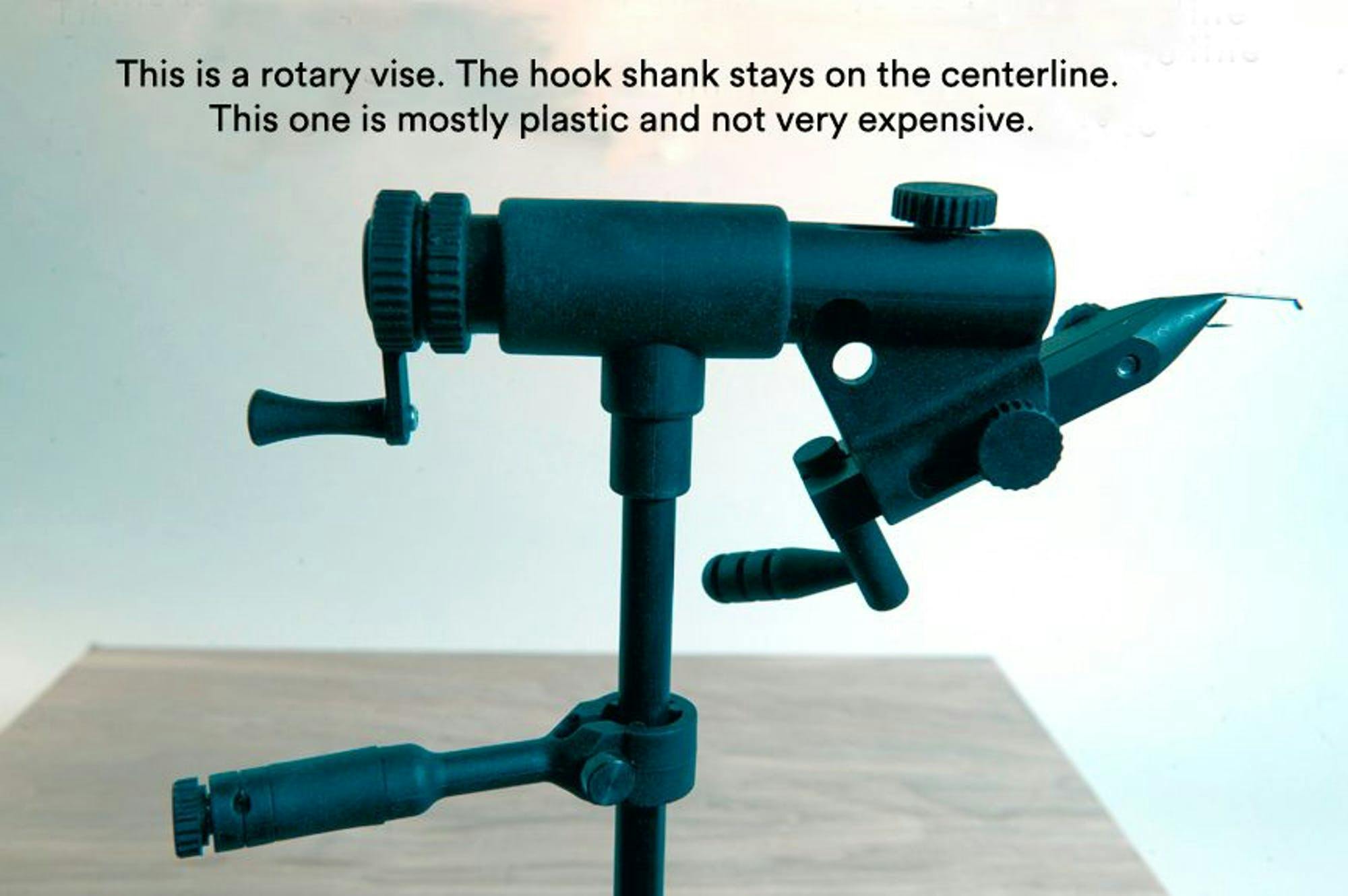 An image of a rotary vise above the plank of wood it's clamped to. It reads "This is a rotary vise. The hook shank stays on the centerline. This one is mostly plastic and not expensive."