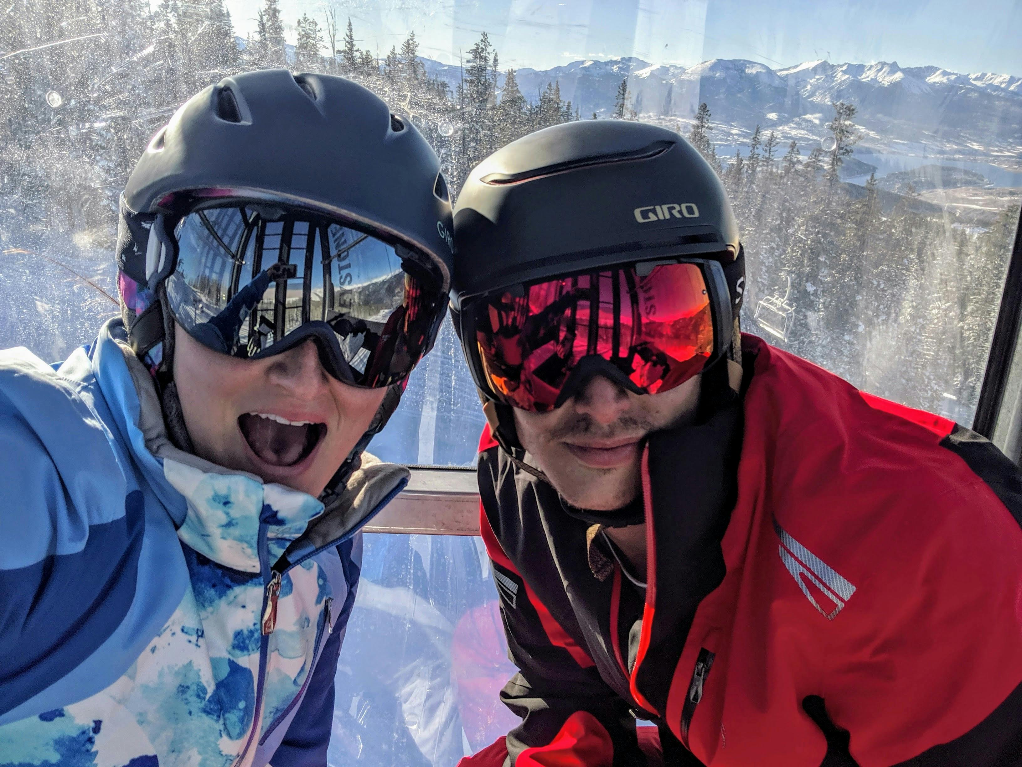 Two skiers in a gondola. They are both wearing helmets and goggles. 