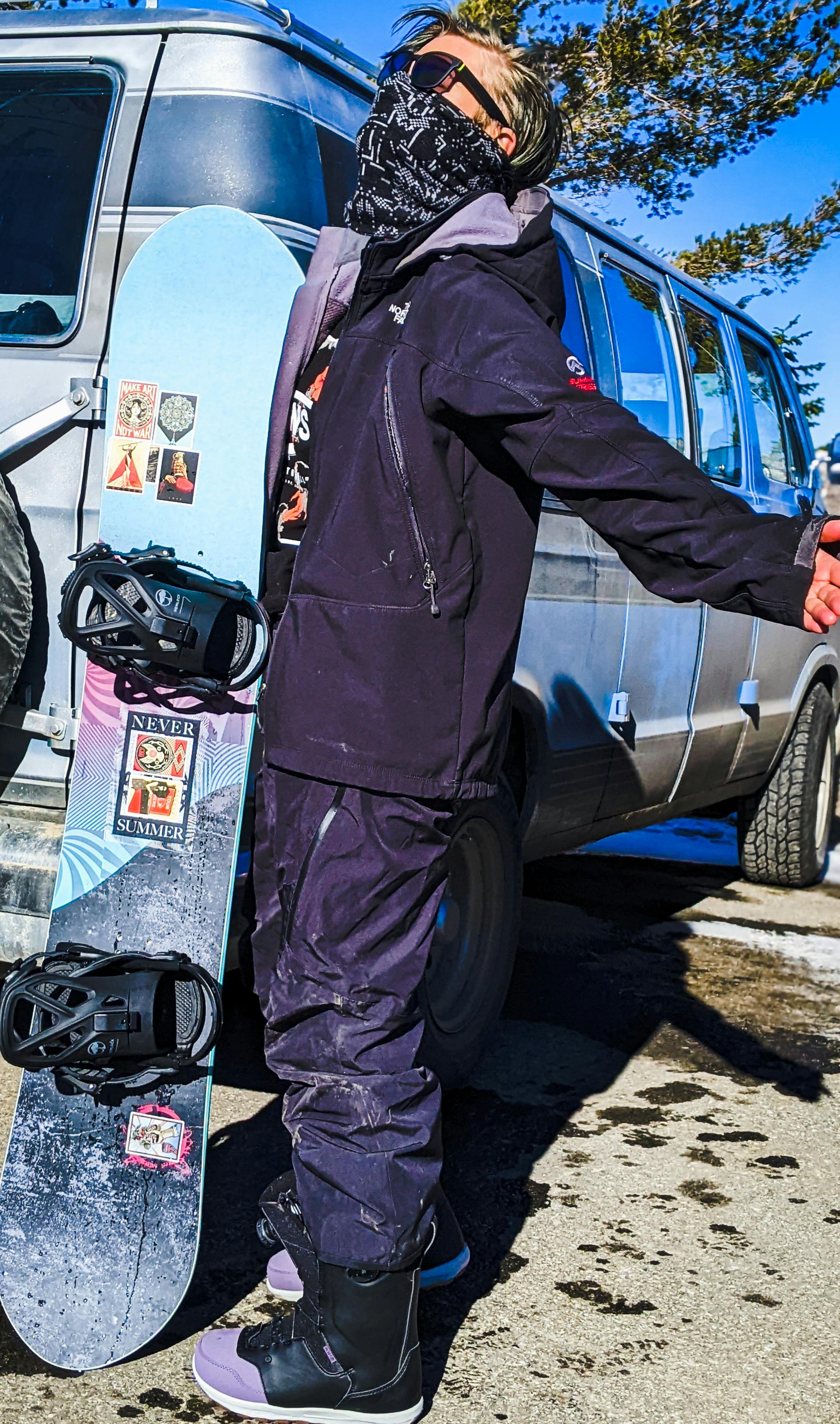 A snowboarder standing with their snowboard near a van. 