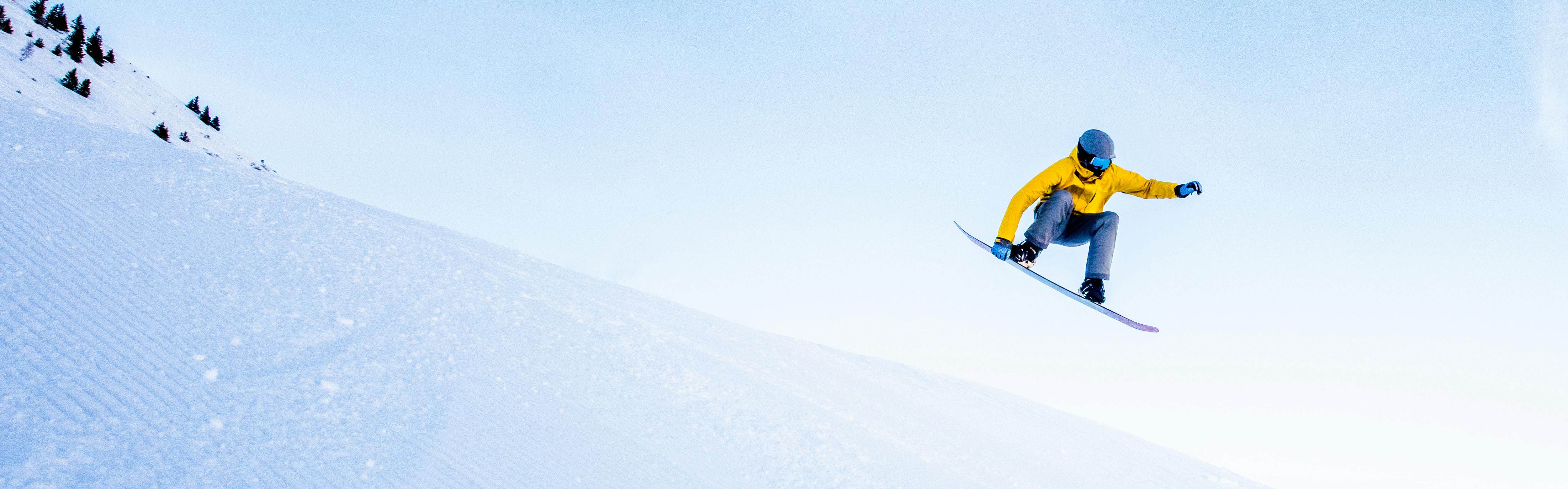 A snowboarder in a yellow jacket does a jump off a groomer trail as he grabs his snowboard. 