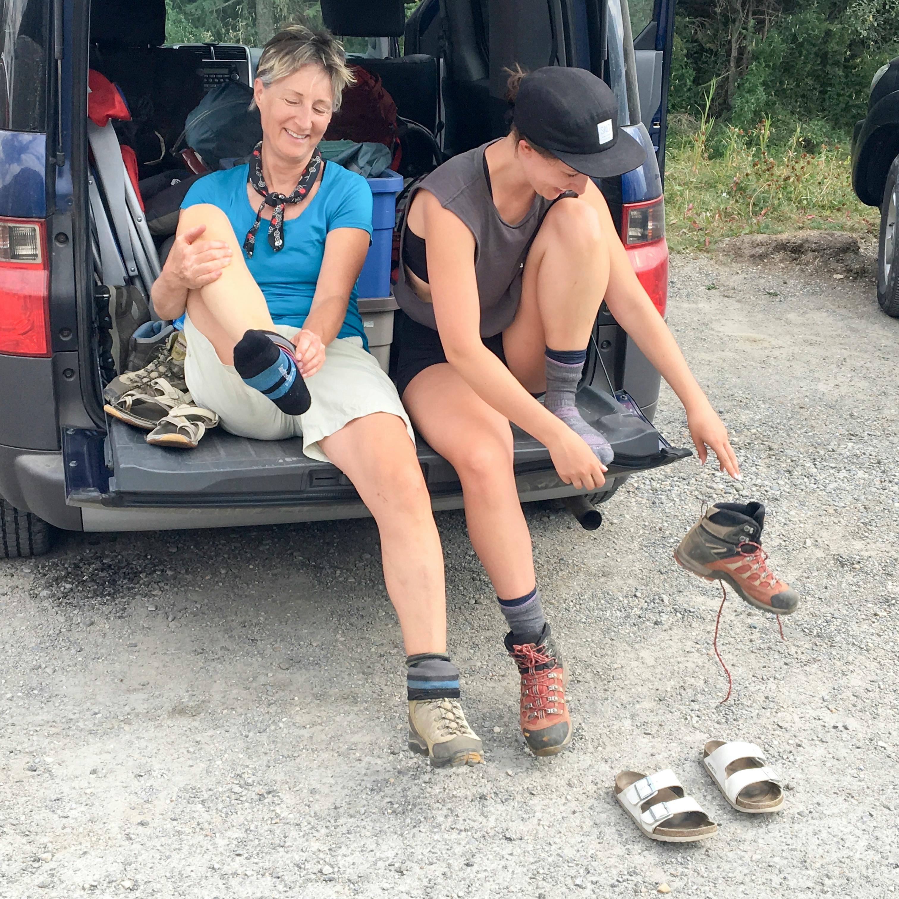 Two women sit in the back of an SUV and remove their hiking boots