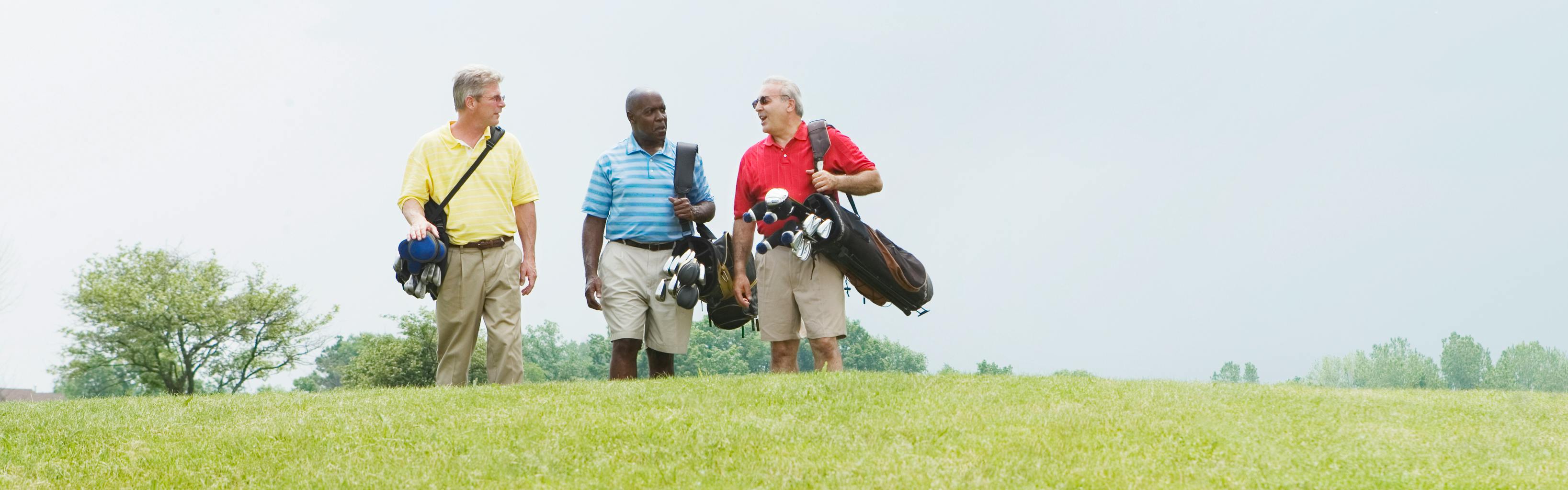 Three older men talk while walking across a golf course. All three carry bags of golf clubs