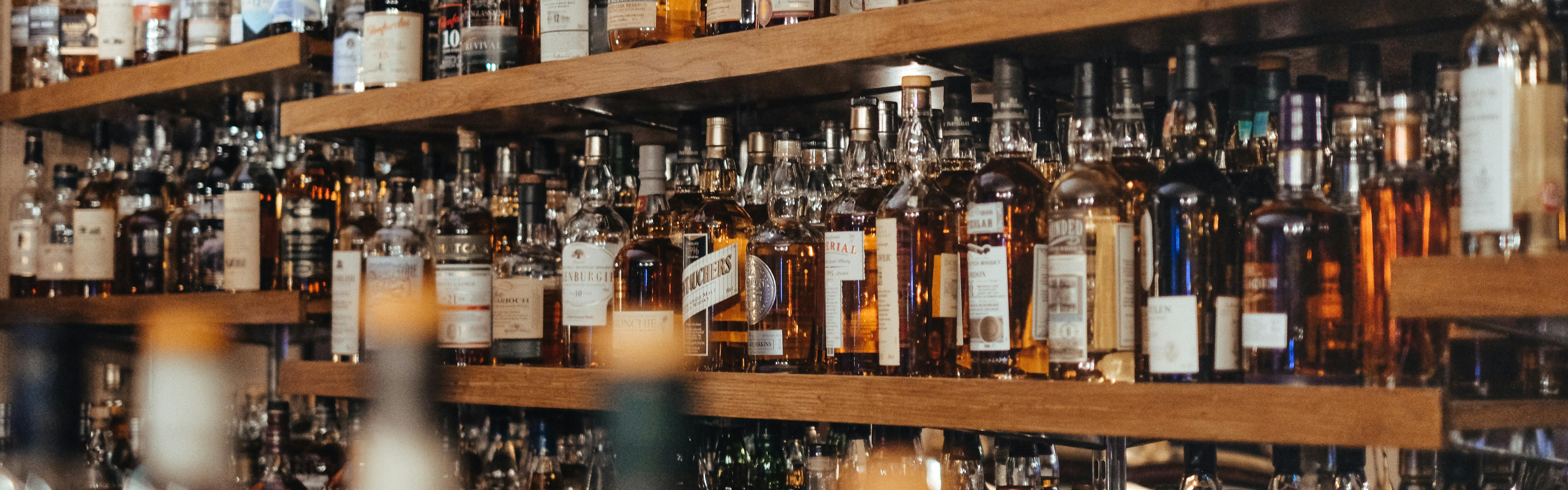 Wooden bar shelves are lined with liquor. Out-of-focus bottlenecks rise in the foreground, partially obscuring the shelves behind. 