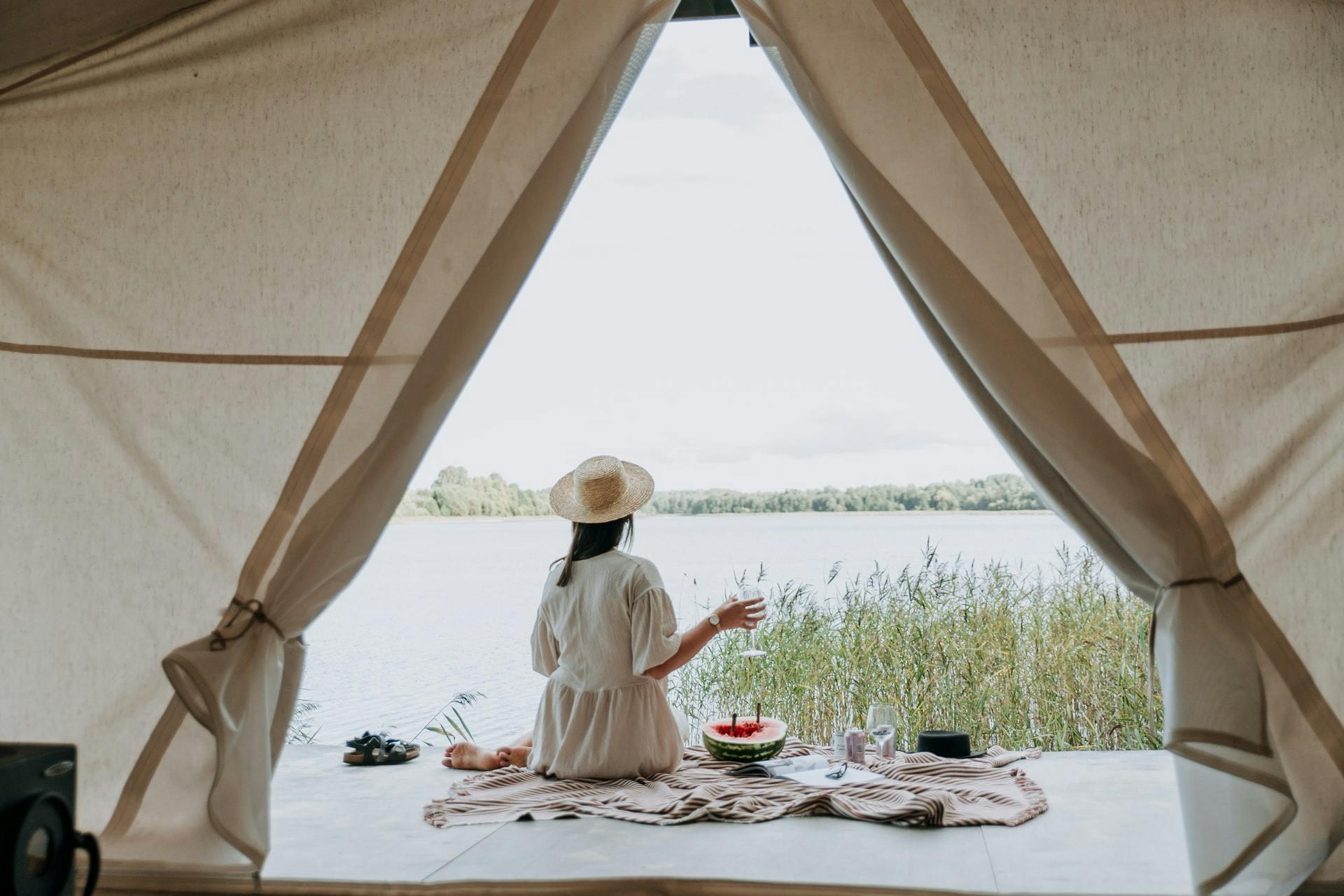 Woman in a hat drinking something and eating some food outside of a tent.