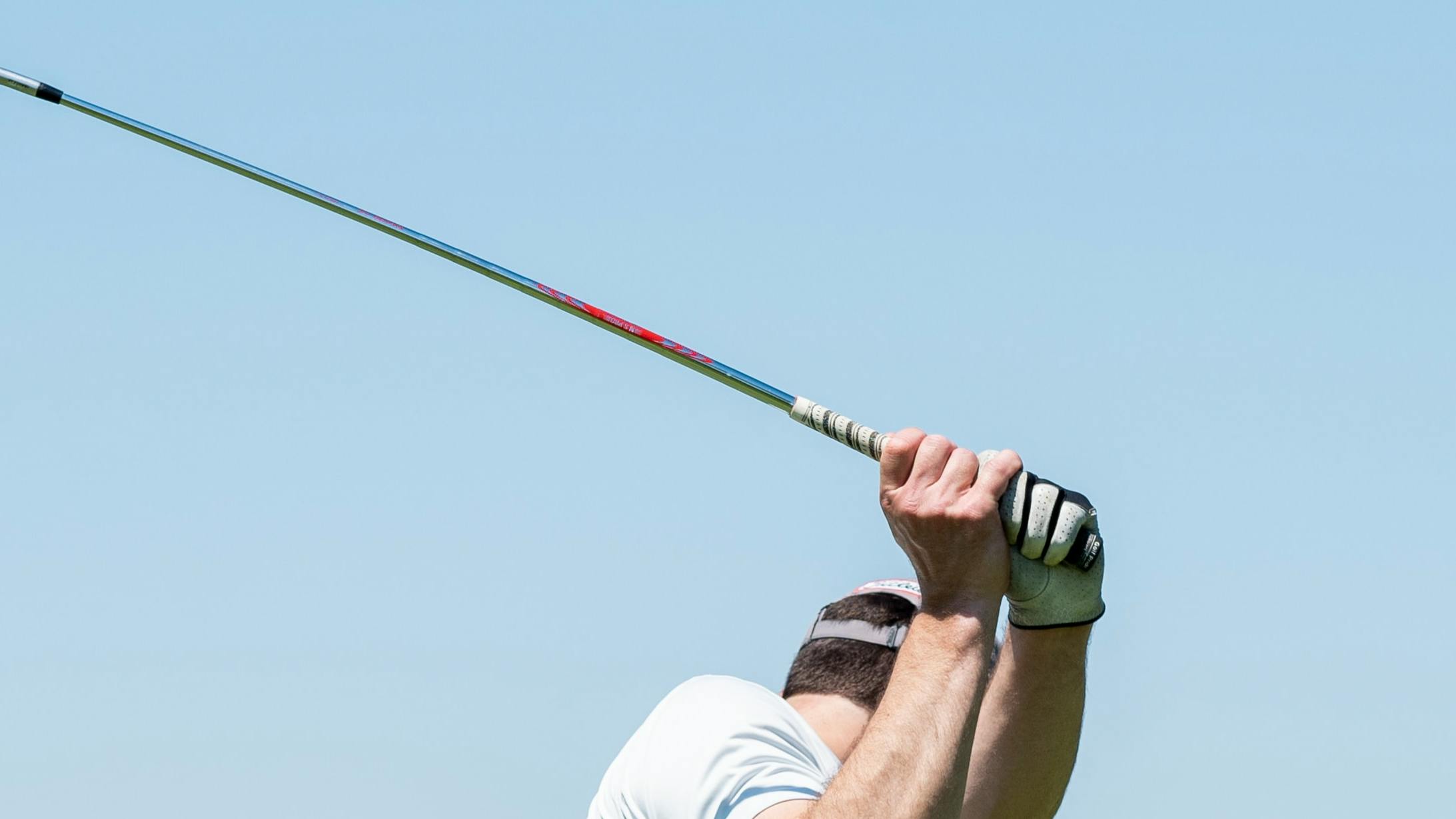 A man swings back his golf club. He is backdropped by a pale blue sky. 