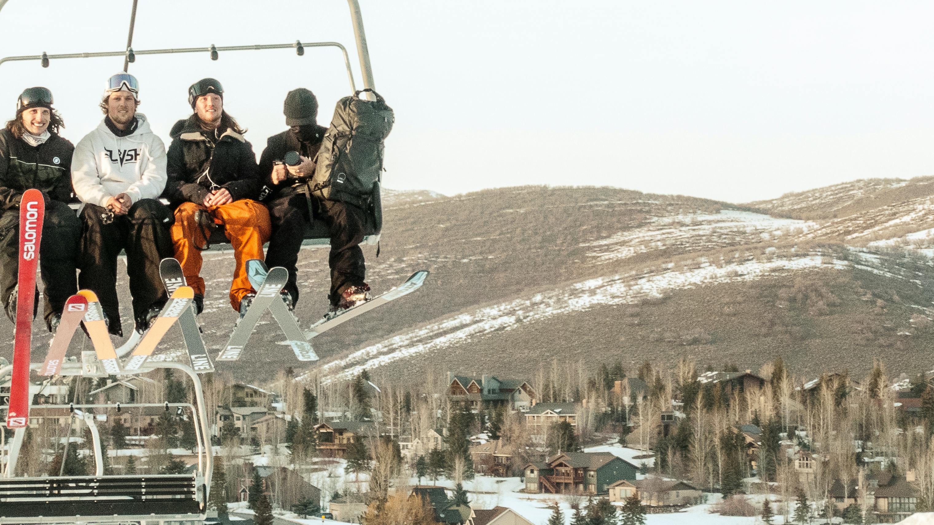 Four skiers on a chairlift. They are all wearing light jackets or hoodies. There is a mountain in the background that has very little snow on it. 
