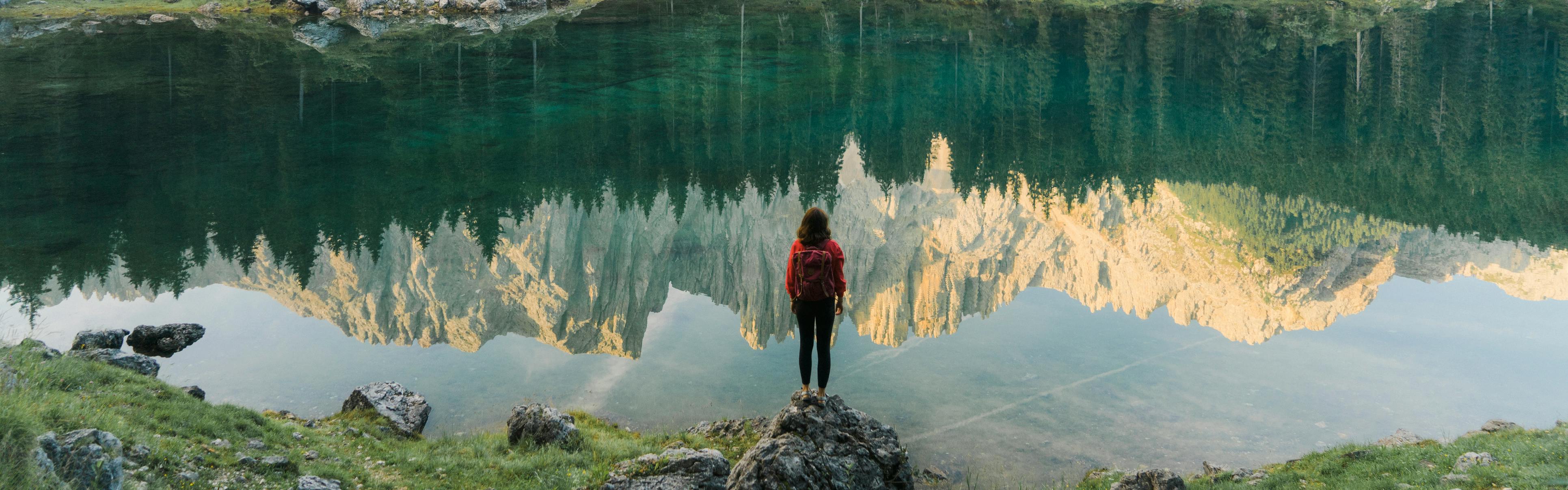 A woman with a backpack stands on a rock and looks out at a clear lake. The lake is reflecting a mountain range