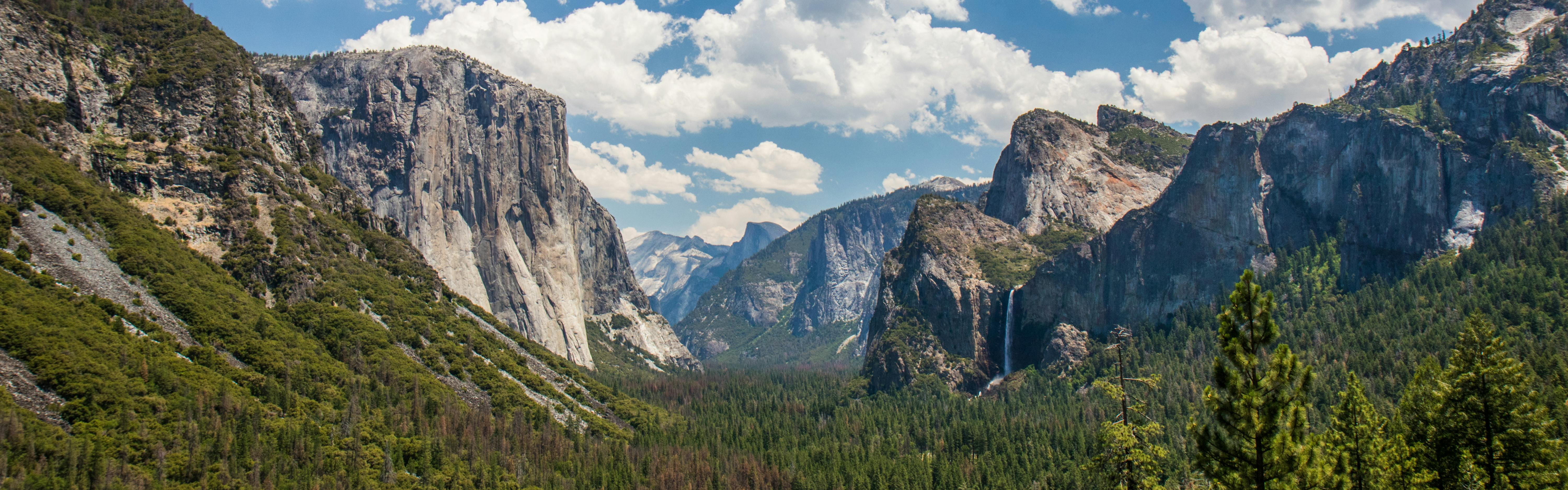 The famous Tunnel View of Yosemite National Park with Half Dome and a waterfall visible on the right side. The landscape is green and lush and the sky above is pale blue with puffy white clouds.