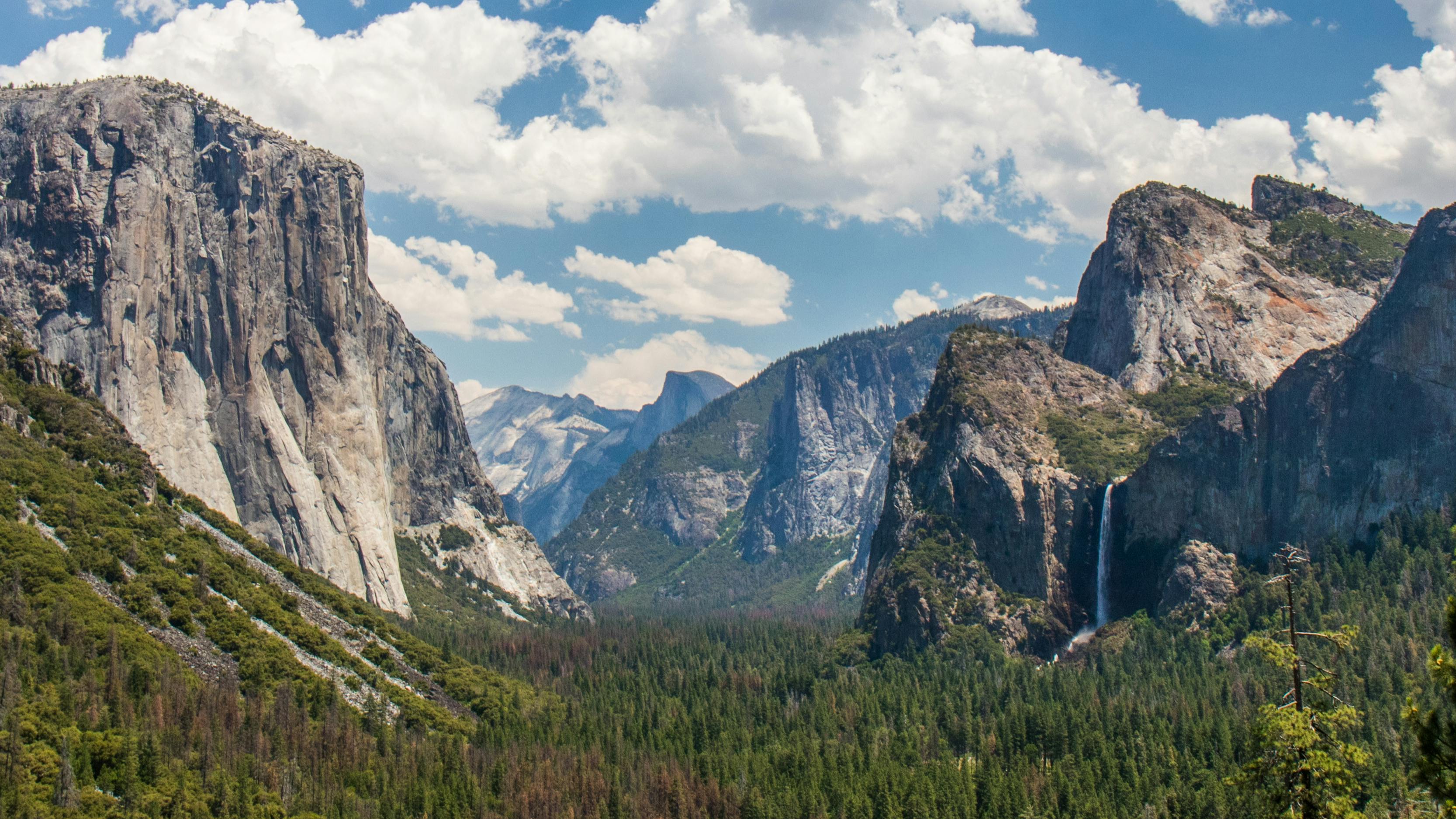 The famous Tunnel View of Yosemite National Park with Half Dome and a waterfall visible on the right side. The landscape is green and lush and the sky above is pale blue with puffy white clouds.