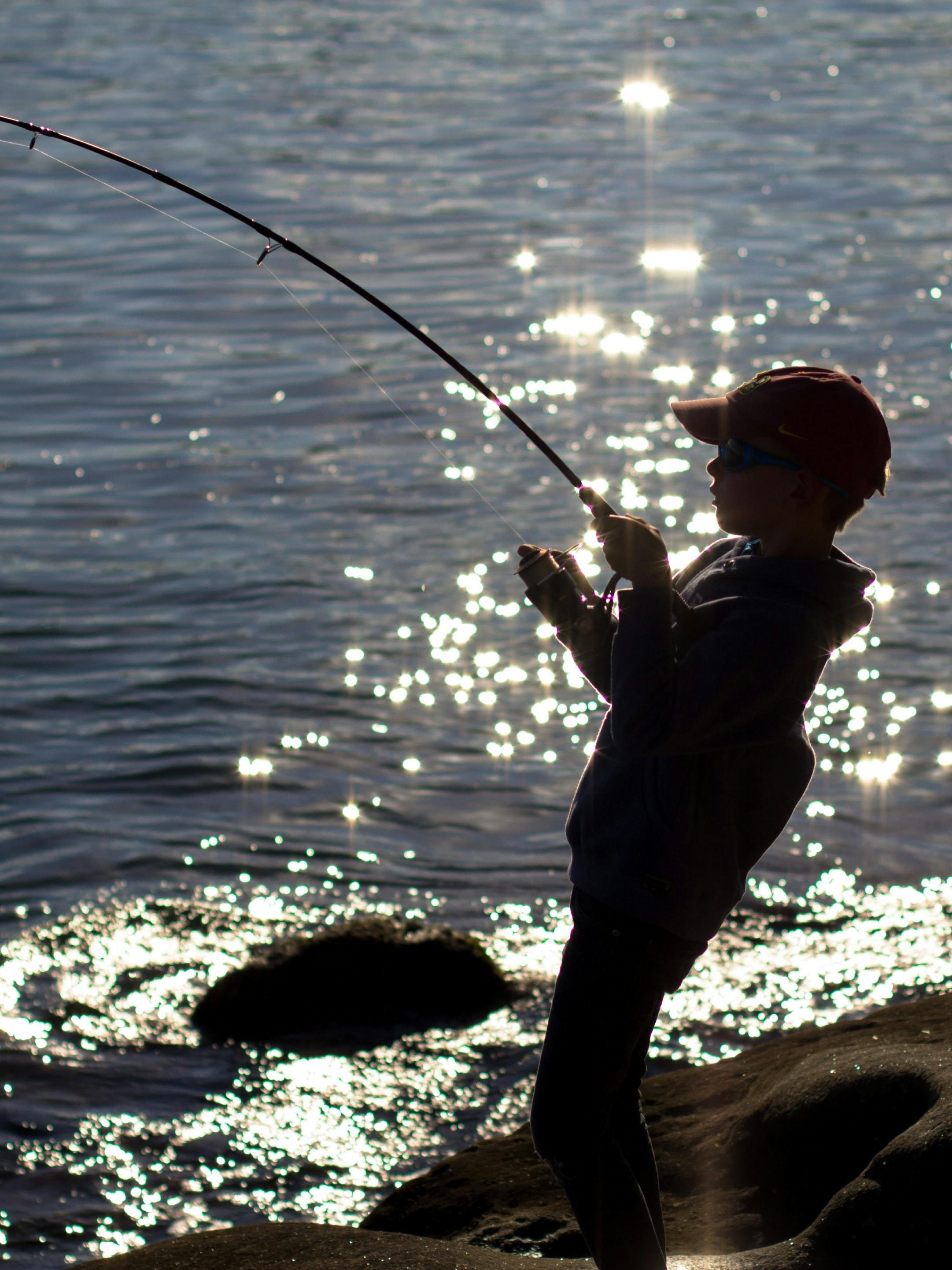 A silhouetted child stands on the banks of a sparking water source, leaning back against a curving fishing rod. 