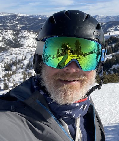 A man takes a selfie with a ski resort visible in the background. He is wearing a helmet and ski gear, including the Smith I/O Mag goggles. 