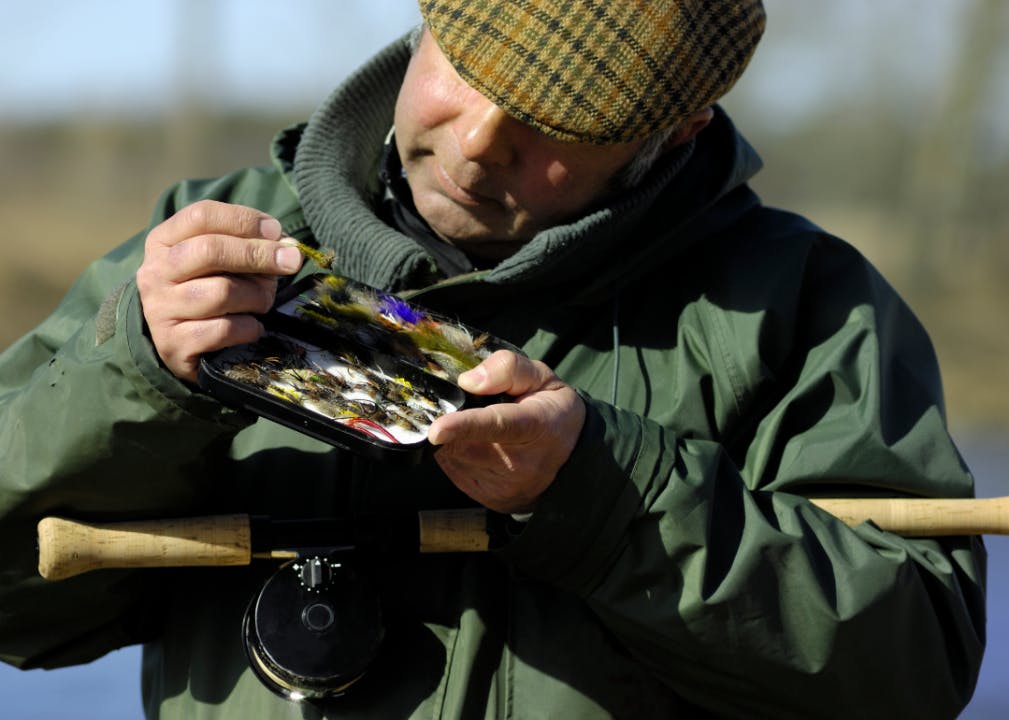 A fly fisherman holding a fly box and examining a fly with a fly rod tucked in the crook of his elbow
