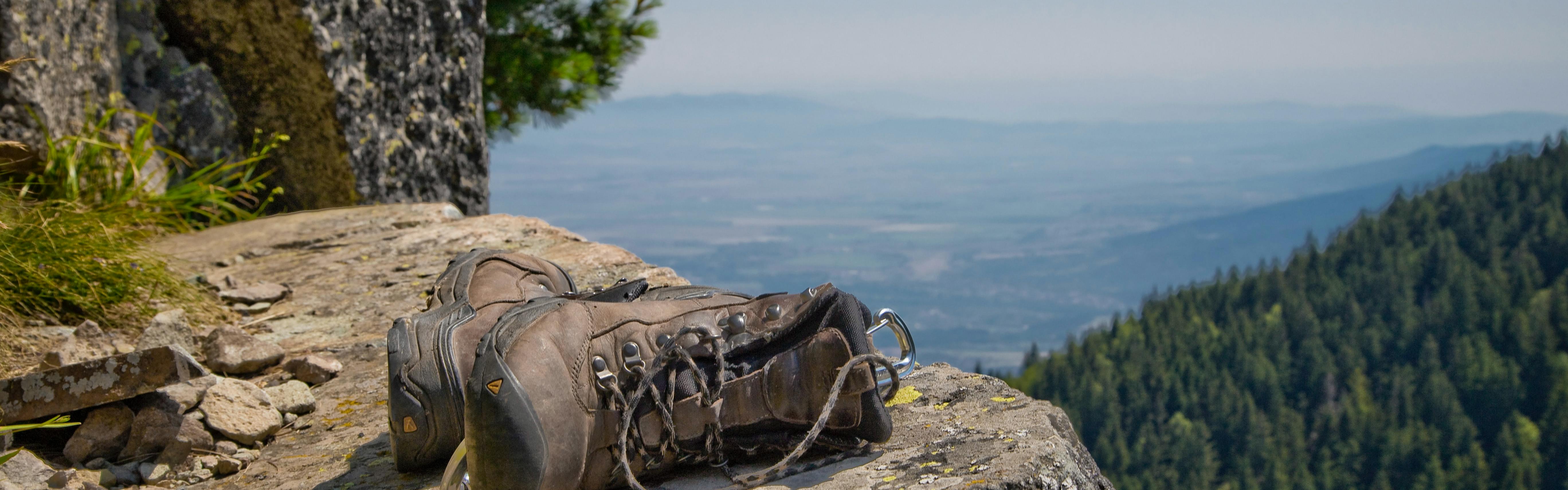 A pair of hiking boots lay sideways on a cliff-side rock with a view of a plain below.