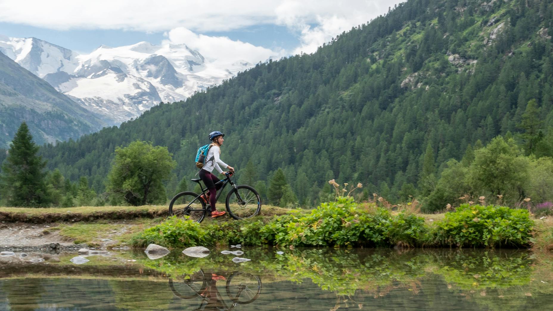 A woman on a mountain bike rides next to a pond with mountains and hills behind her