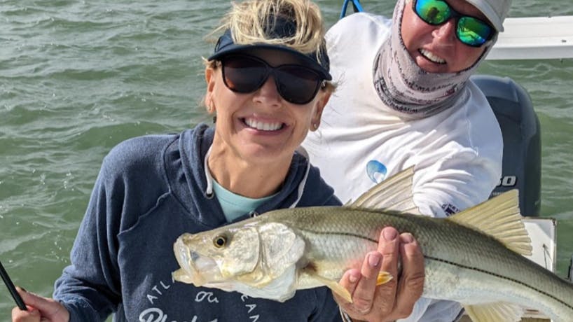 A man and a woman smile at the camera as they hold up a snook. They are on a boat and the woman is holding a fishing pole. 