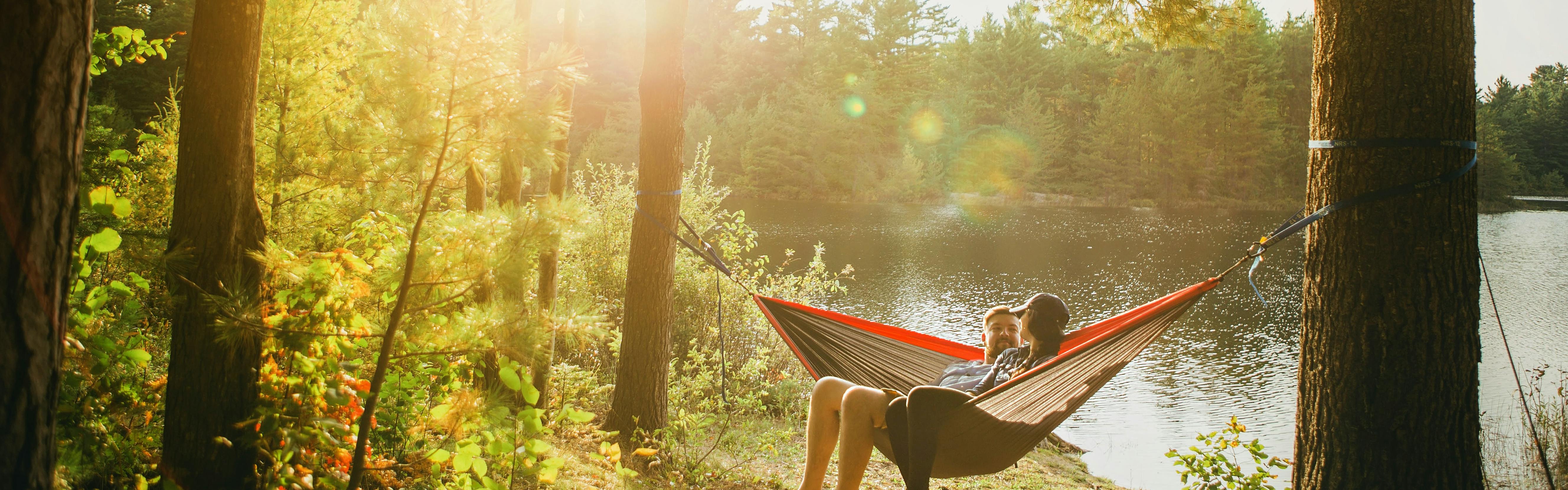 Two people sit in a hammock next to a lake. The hammock is set up between two conifers. The sun filters through the foliage.