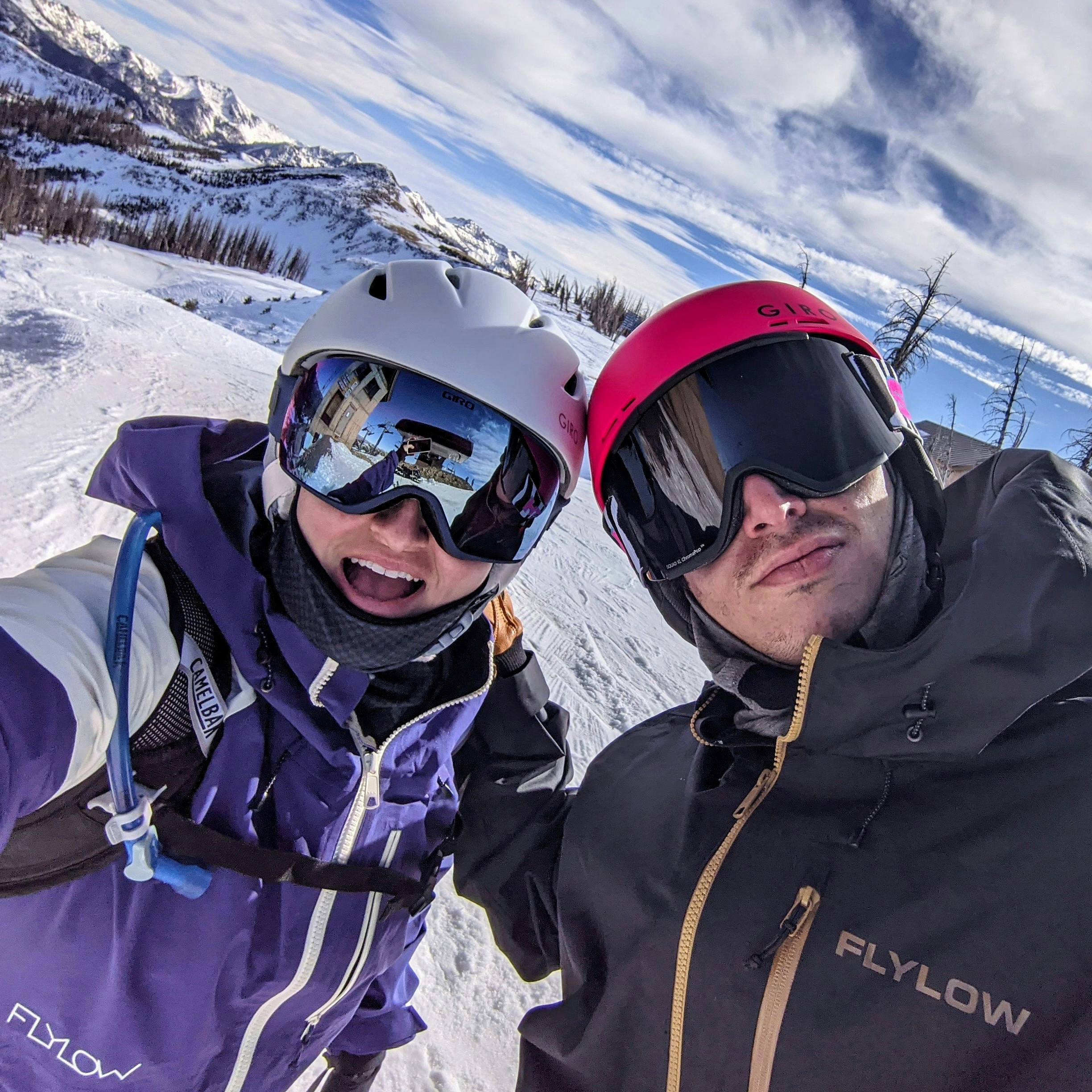 A skier taking a selfie with another skier. They are both wearing helmets and smiling. There is a snowy ski mountain in the background. 