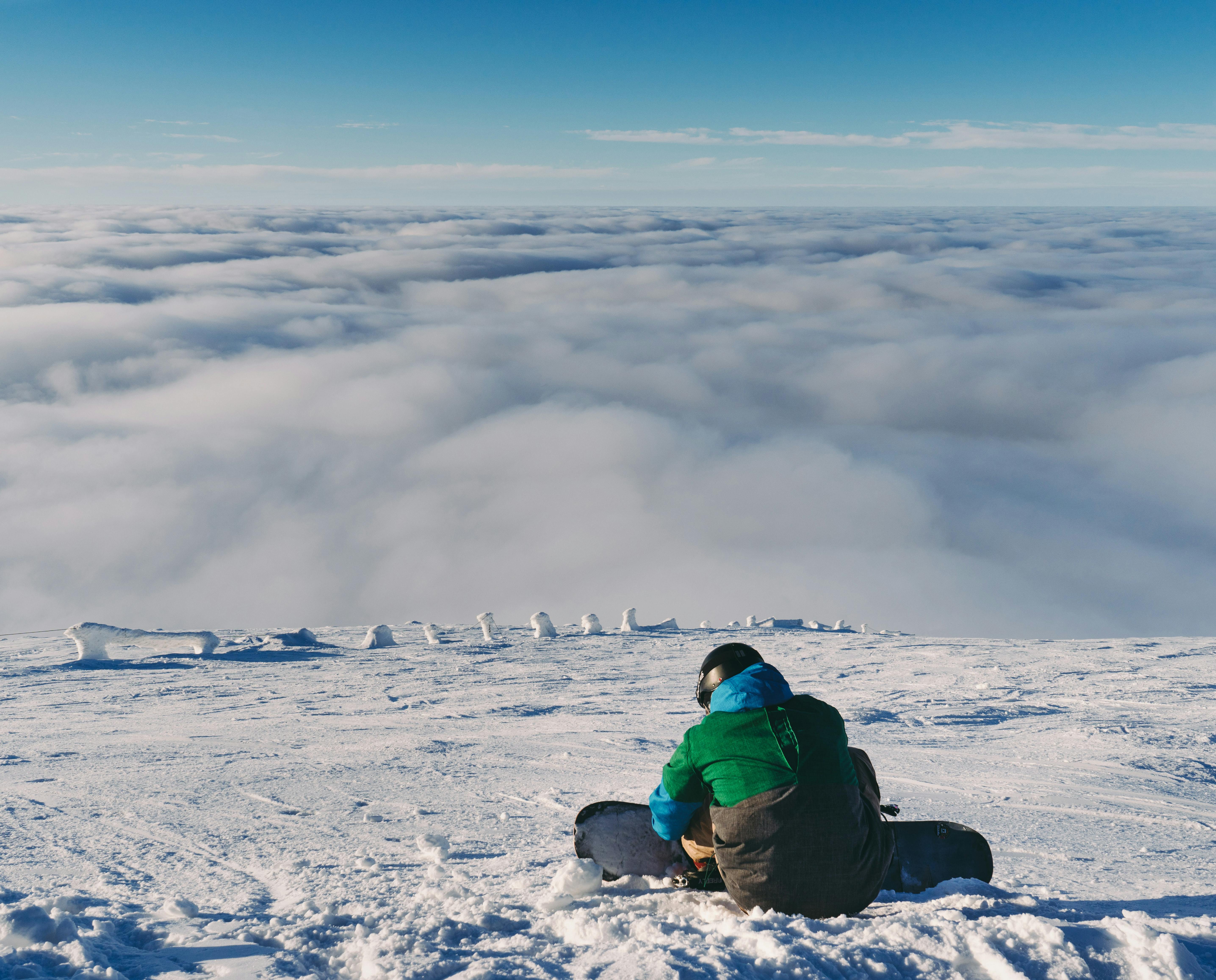 A snowboarder sits by themselves on a flat area with a view of the clouds stretching before them.