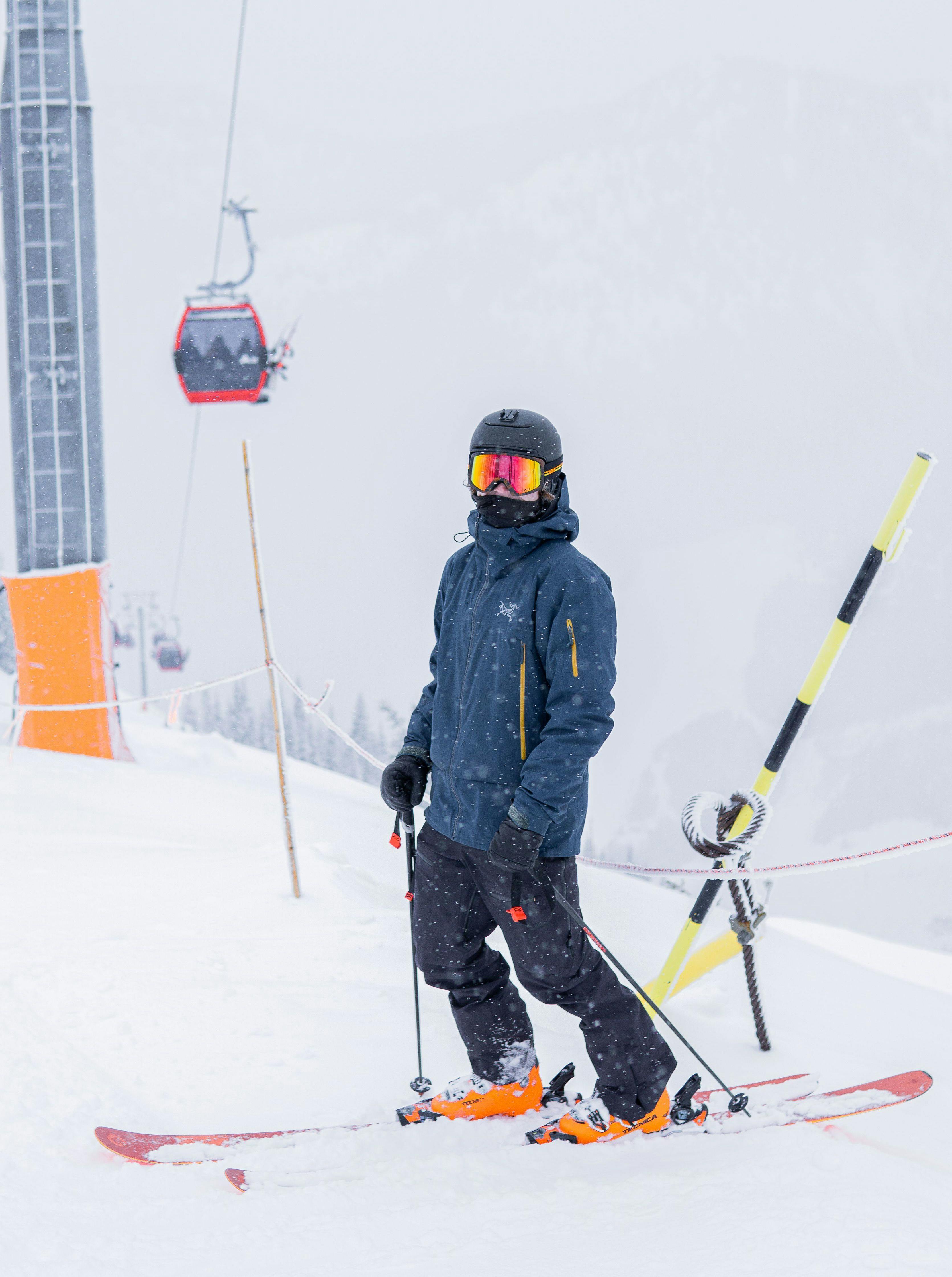 Someone looks at the camera while on skis, wearing a helmet and goggles. Behind them, we can see a gondola run. 
