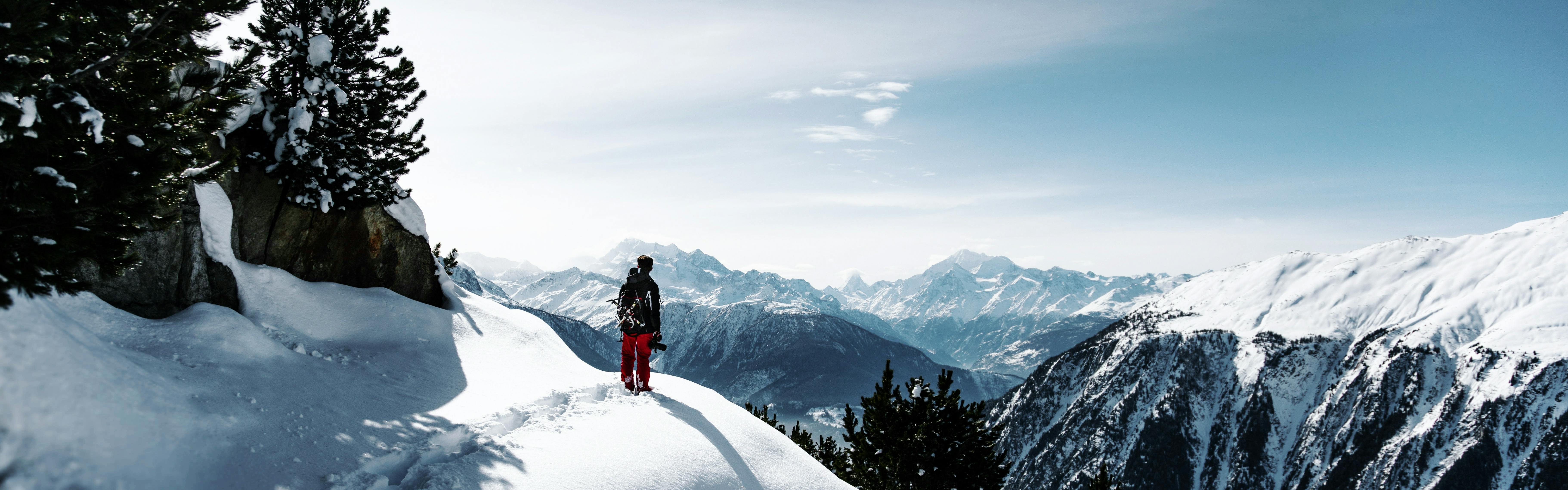 A man stands on a snowy trail that overlooks snowy peaks.