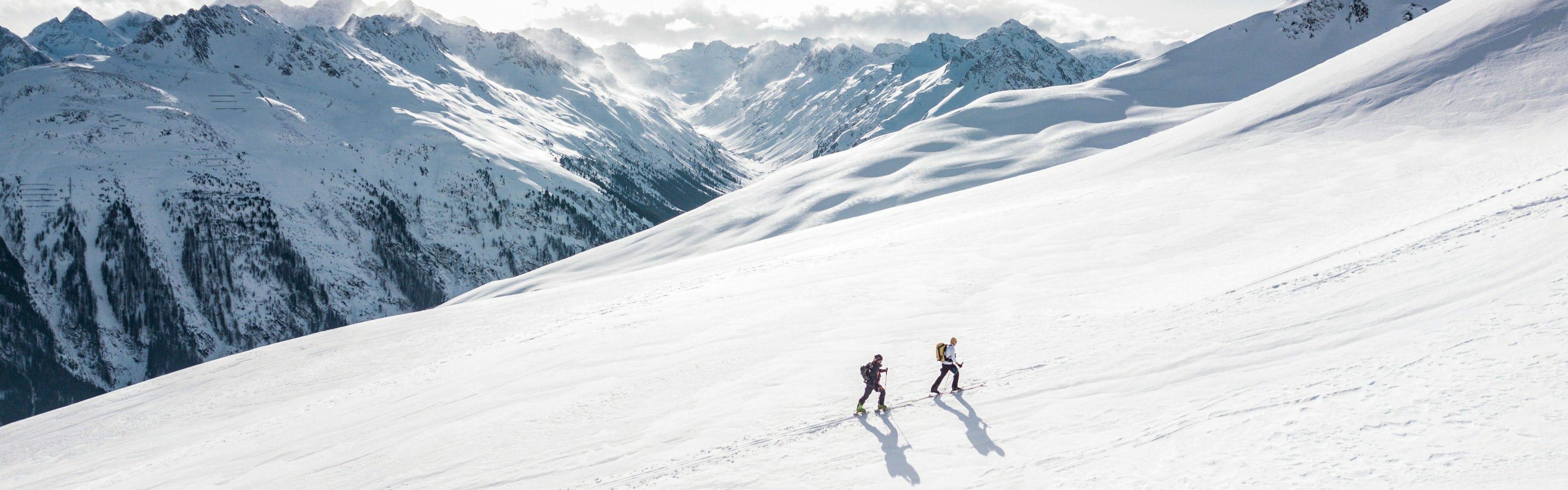 Two skiers walking up a very snowy hill on skis. There are many snowy mountains in the background.